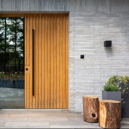 GENERAL HELP Sp.z.o.o - Nowoczesne wejście do domu z drewnianymi drzwiami z pionowymi panelami, długim czarnym uchwytem i szklanym panelem bocznym, otoczone elewacją z szarej cegły, oświetleniem zewnętrznym...