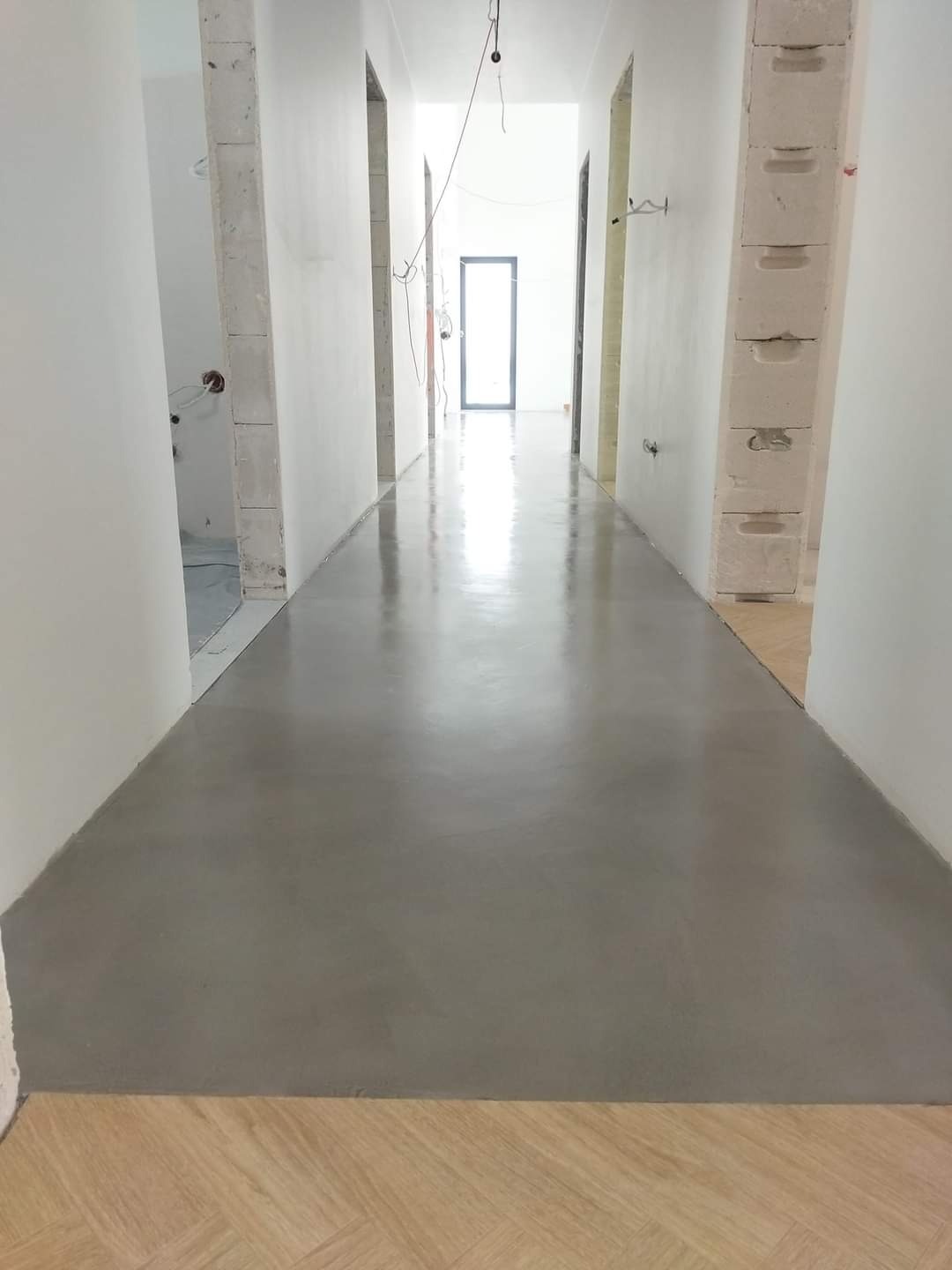 Wnętrze korytarza w trakcie prac wykończeniowych, widoczna wylewka samopoziomująca na podłodze i surowe ściany z pustaków, częściowo wykończone parkietem u dołu kadru.