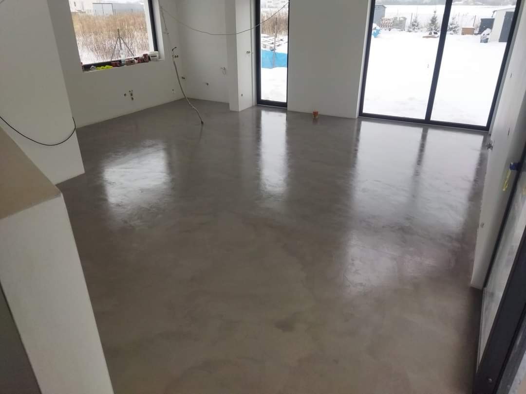 Wnętrze w stanie deweloperskim z wylewką samopoziomującą na podłodze i dużymi oknami z widokiem na zimowy krajobraz.