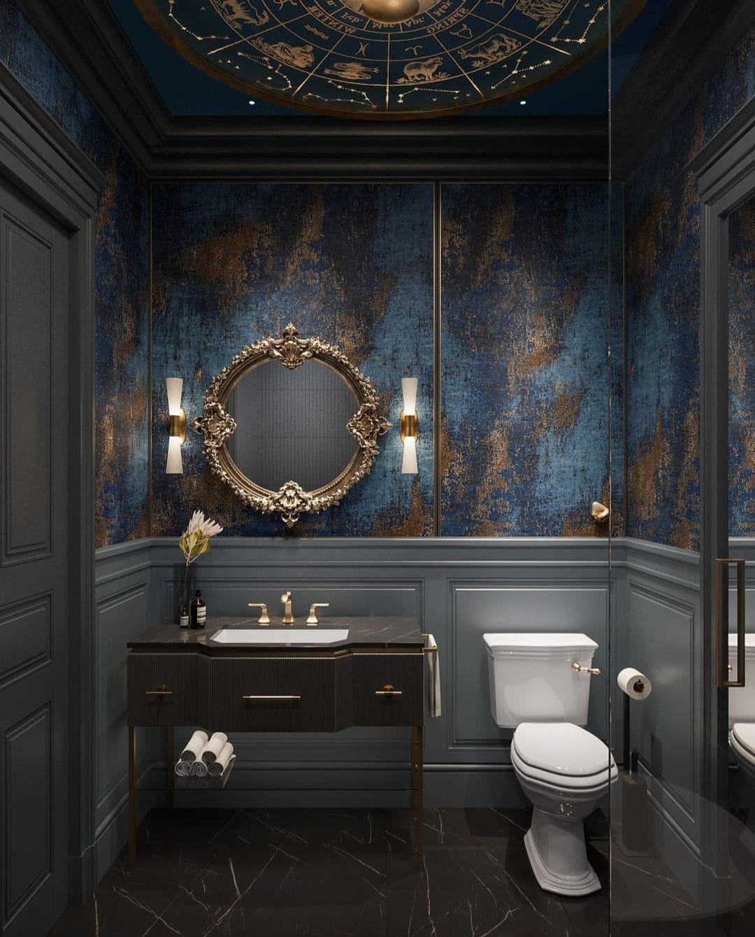 Elegancka, ciemna łazienka z marmurową podłogą, umywalką na złotych nóżkach, ozdobnym lustrem w złotej ramie i tapetą w odcieniach niebieskiego i złota, sufit zdobiony motywami astrologicznymi.