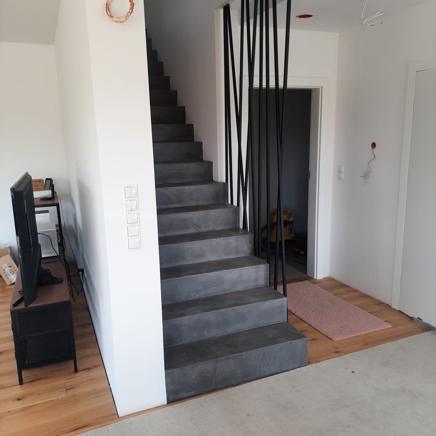 Betonowe schody z minimalistyczną czarną balustradą z prętów w nowoczesnym wnętrzu, widoczny fragment salonu z telewizorem i wykończeniem podłogi w jasnym drewnie.