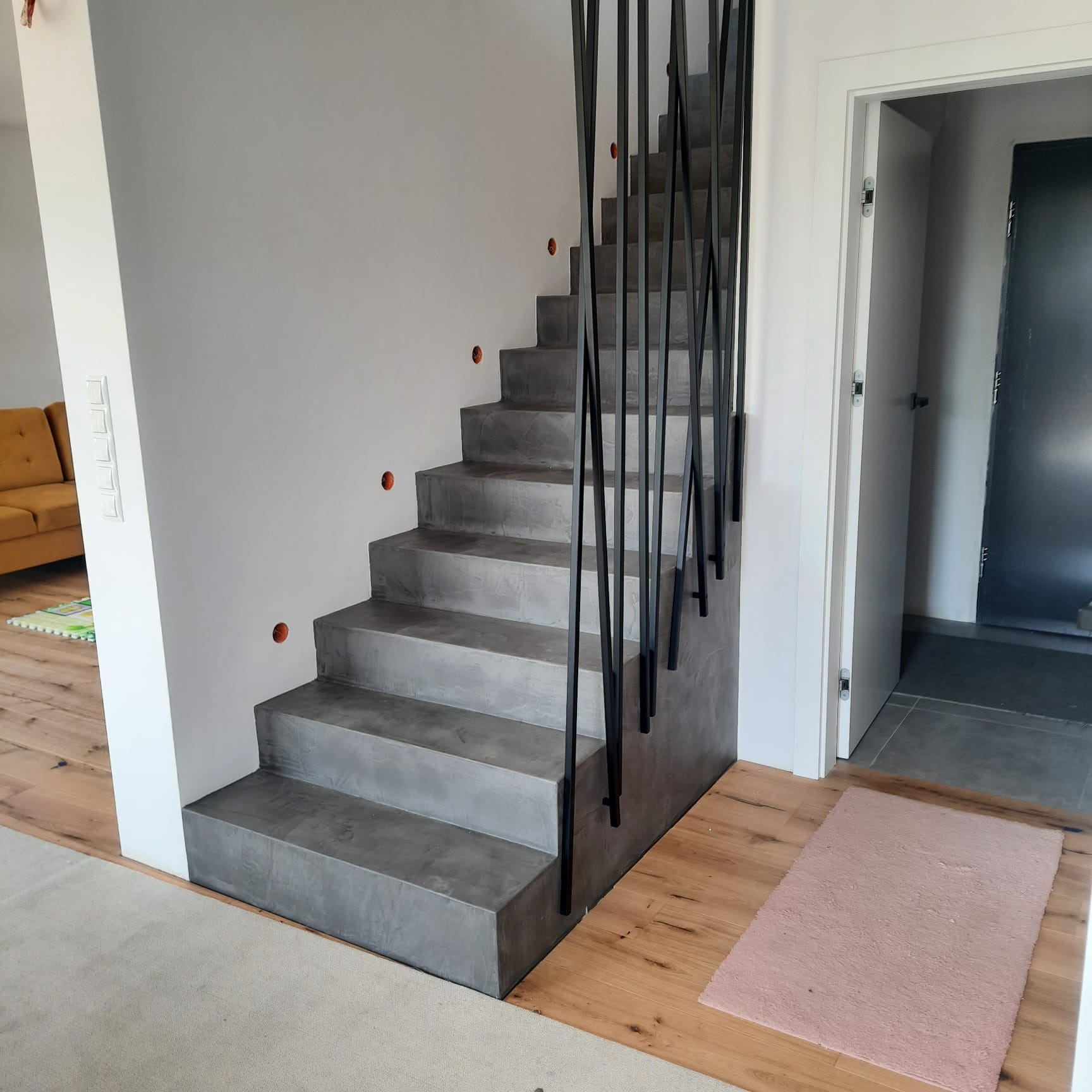 Betonowe schody z minimalistyczną czarną balustradą w nowoczesnym wnętrzu domu, widoczne otwory na oświetlenie w ścianie, jasna podłoga i częściowo widoczny żółty narożnik.