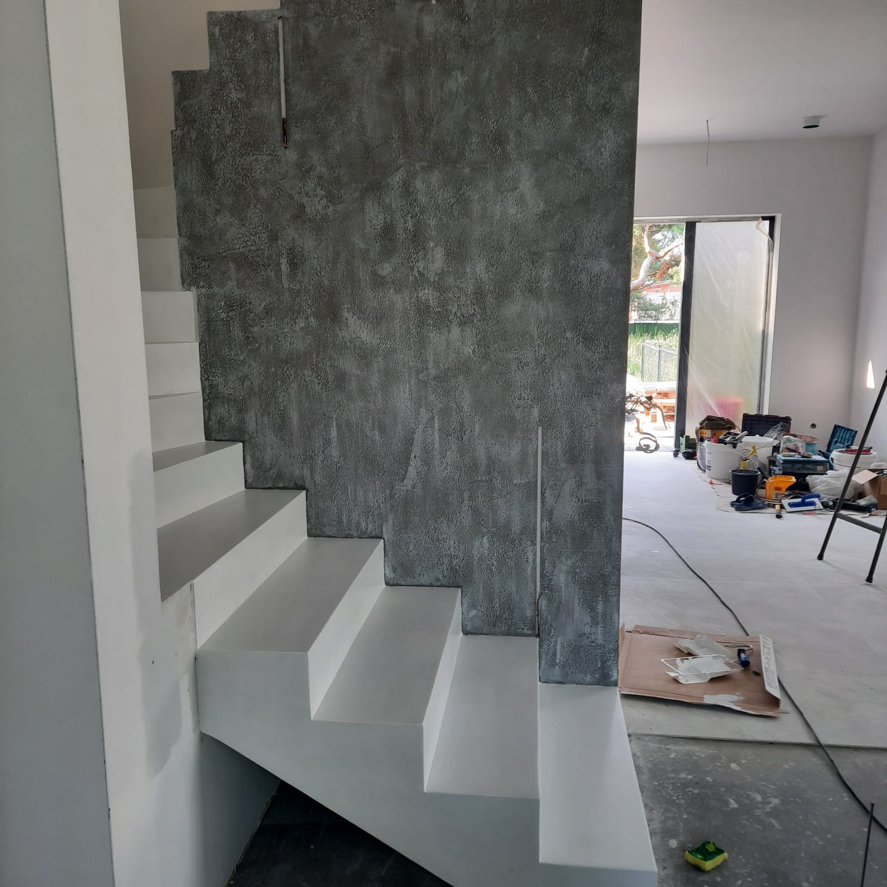 Wnętrze w trakcie prac wykończeniowych: białe schody z szarą ścianą o dekoracyjnej fakturze, w tle widoczne narzędzia i materiały budowlane.