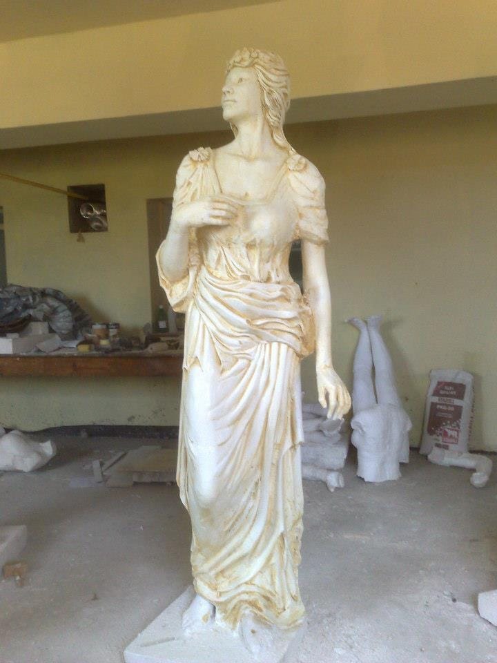 Biała, marmurowa statua kobiety w drapowanej szacie, ustawiona w warsztacie rzeźbiarskim z widocznymi narzędziami i elementami w trakcie obróbki.