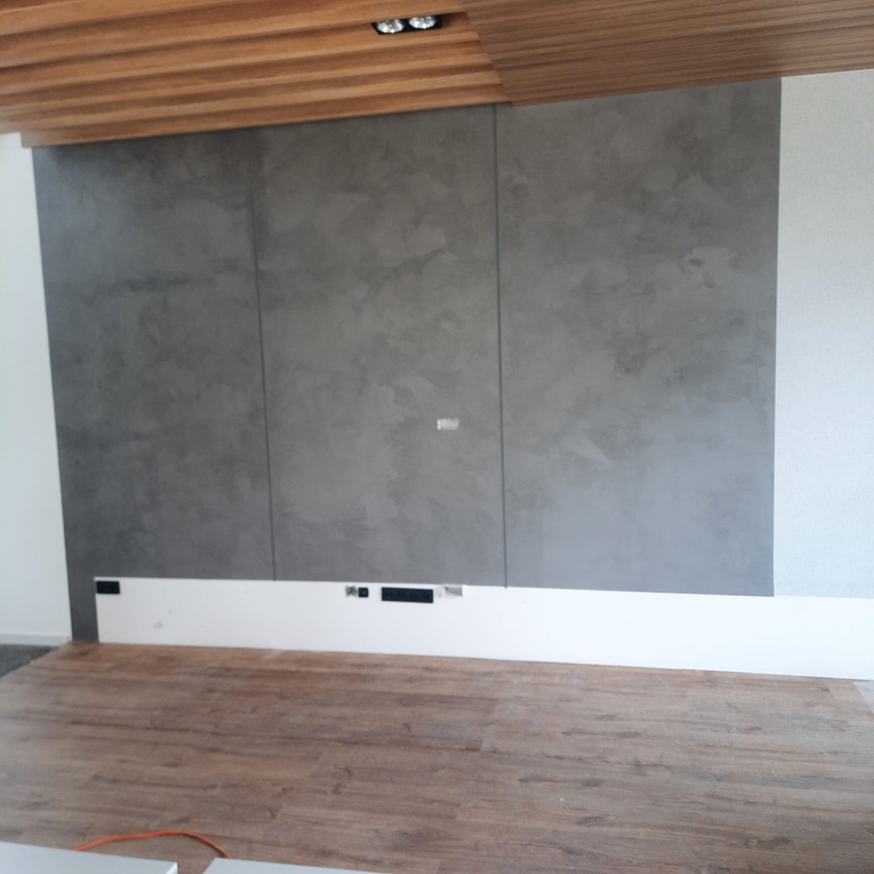 Ściana z efektem dekoracyjnym w odcieniach szarości, podzielona na panele, z drewnianym sufitem i panelową listwą przypodłogową z otworami na kable.