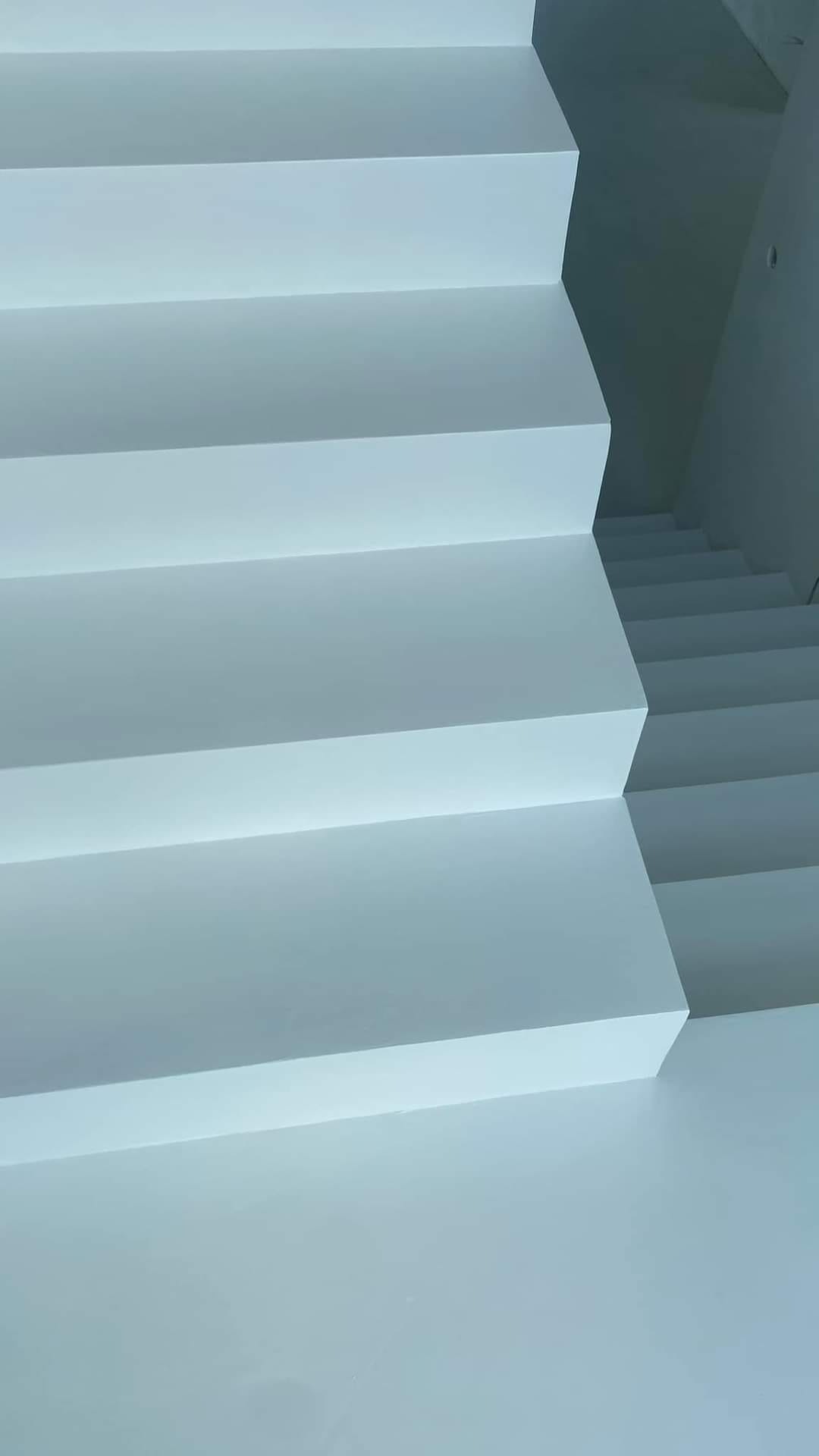 Minimalistyczne, białe schody wewnętrzne o geometrycznym designie, z widocznymi stopniami i przestrzenią pod schodami.