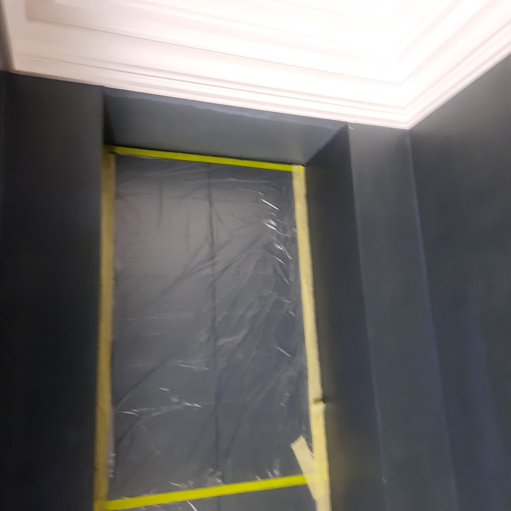 Świeżo pomalowana na ciemny grafit ściana z białym sufitem, zabezpieczona folią i taśmą malarską w trakcie prac wykończeniowych.