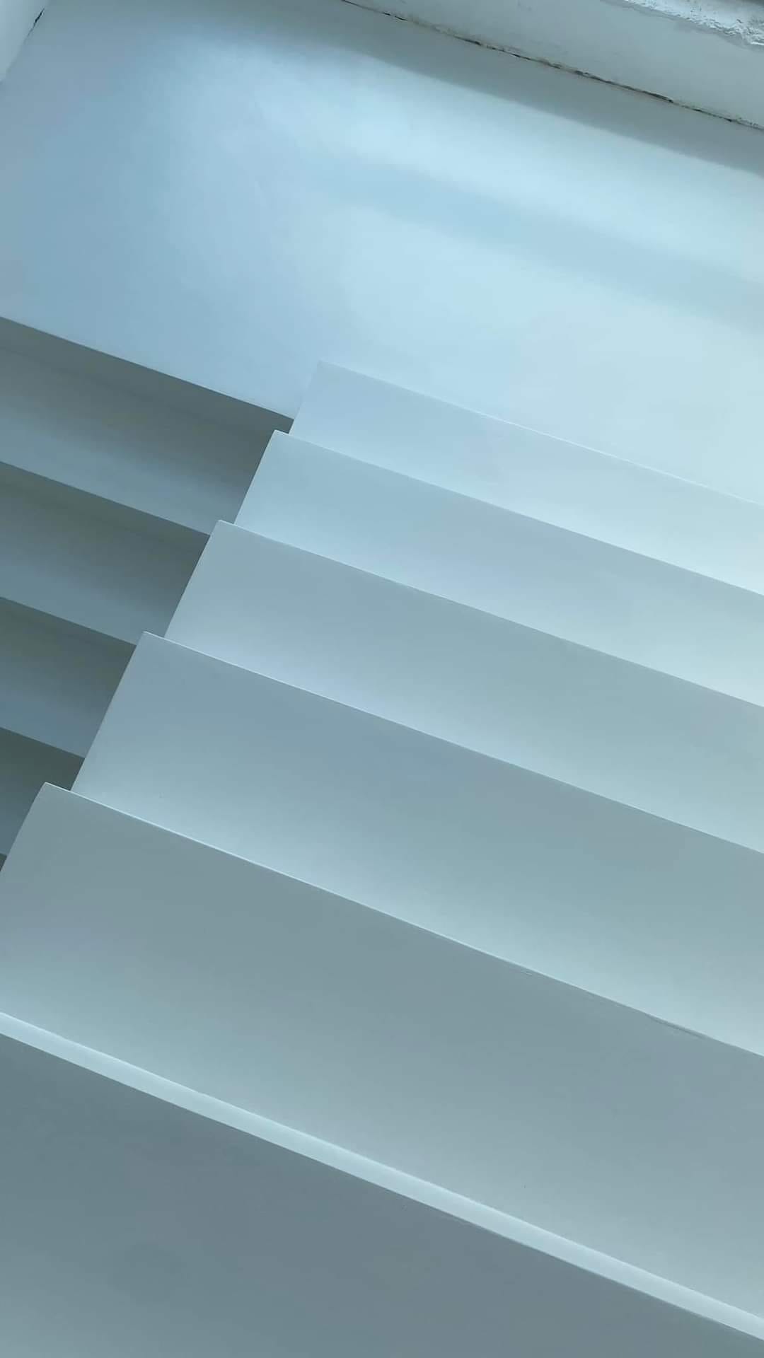 Minimalistyczne, białe schody wewnętrzne prowadzące do góry, oświetlone od góry, tworzące geometryczny wzór cieni.