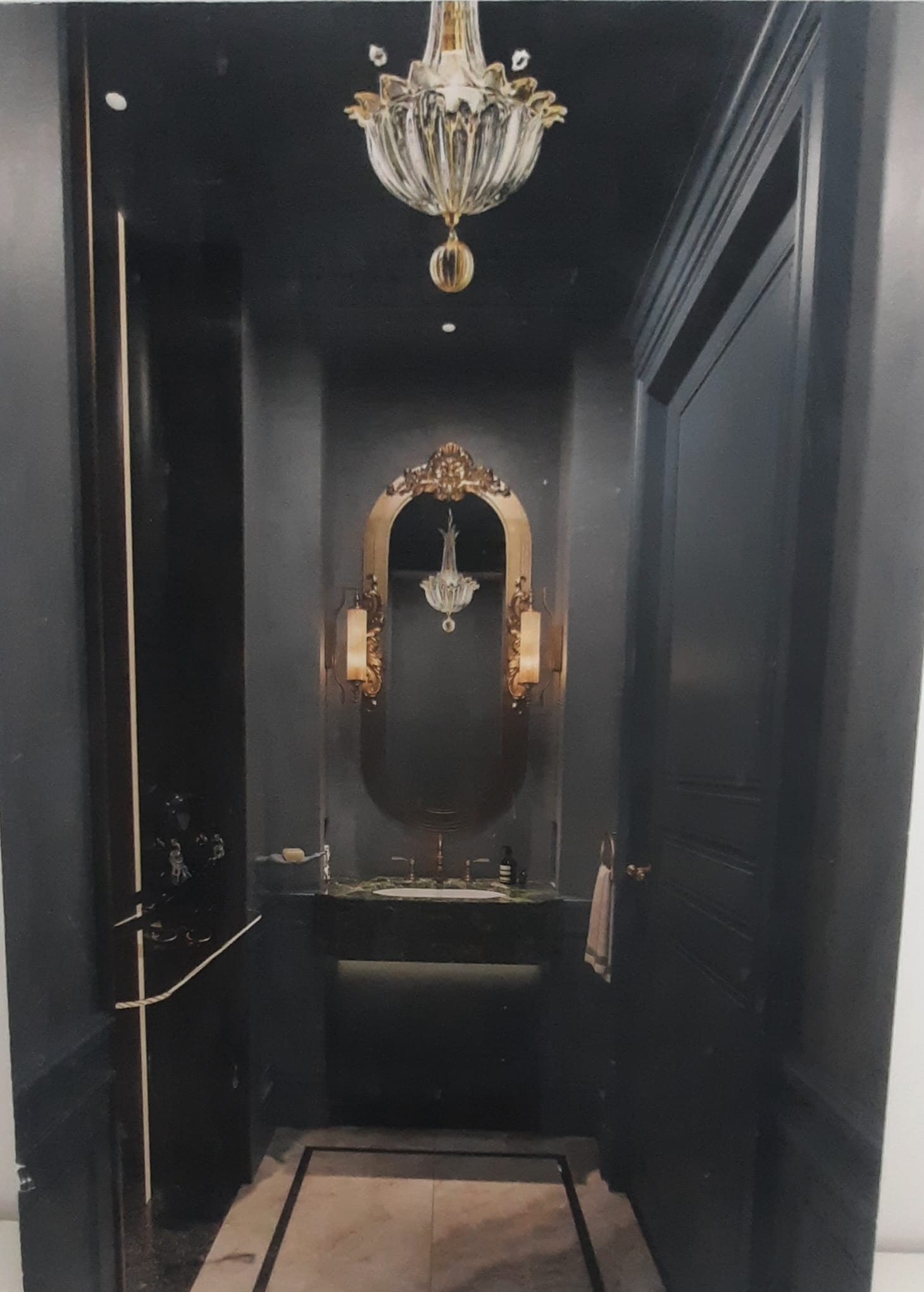 Elegancka łazienka w ciemnej tonacji z marmurową podłogą, ozdobnym lustrem w złotej ramie i kryształowym żyrandolem.