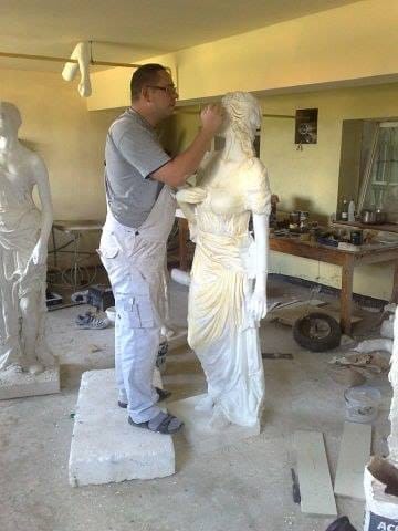 Artysta w szarym t-shircie i białych spodniach roboczych retuszuje gipsową statuę kobiety w pracowni, widoczne inne rzeźby i narzędzia.