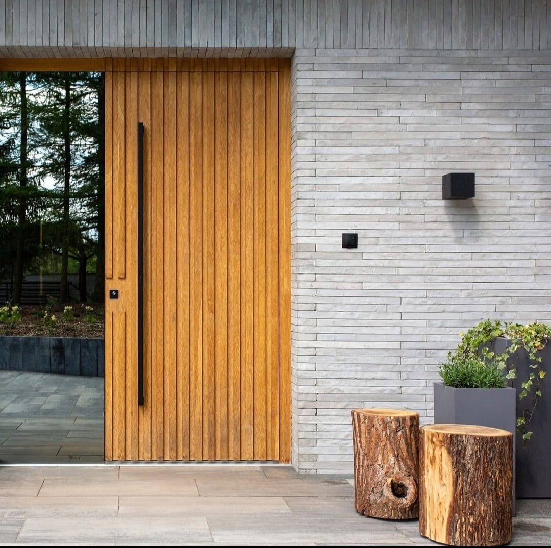 Nowoczesne wejście do domu z drewnianymi drzwiami z pionowymi panelami, długim czarnym uchwytem i szklanym panelem bocznym, otoczone elewacją z szarej cegły, oświetleniem zewnętrznym...