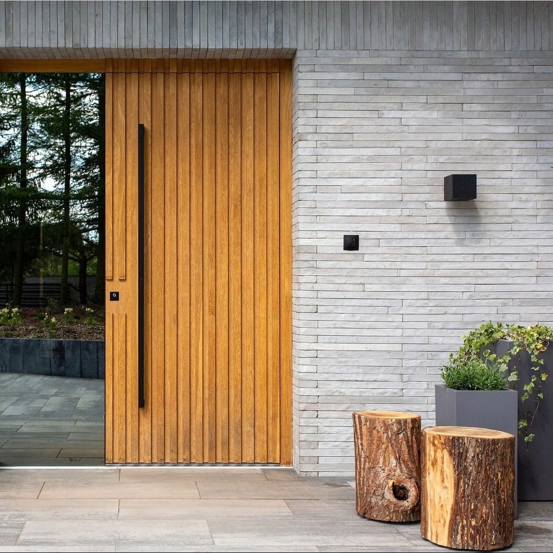 Nowoczesne wejście do domu z drewnianymi drzwiami z pionowymi panelami i długim, czarnym uchwytem, ściana wykończona szarym kamieniem, oświetlenie zewnętrzne w formie czarnych kostek, dwa drewniane...