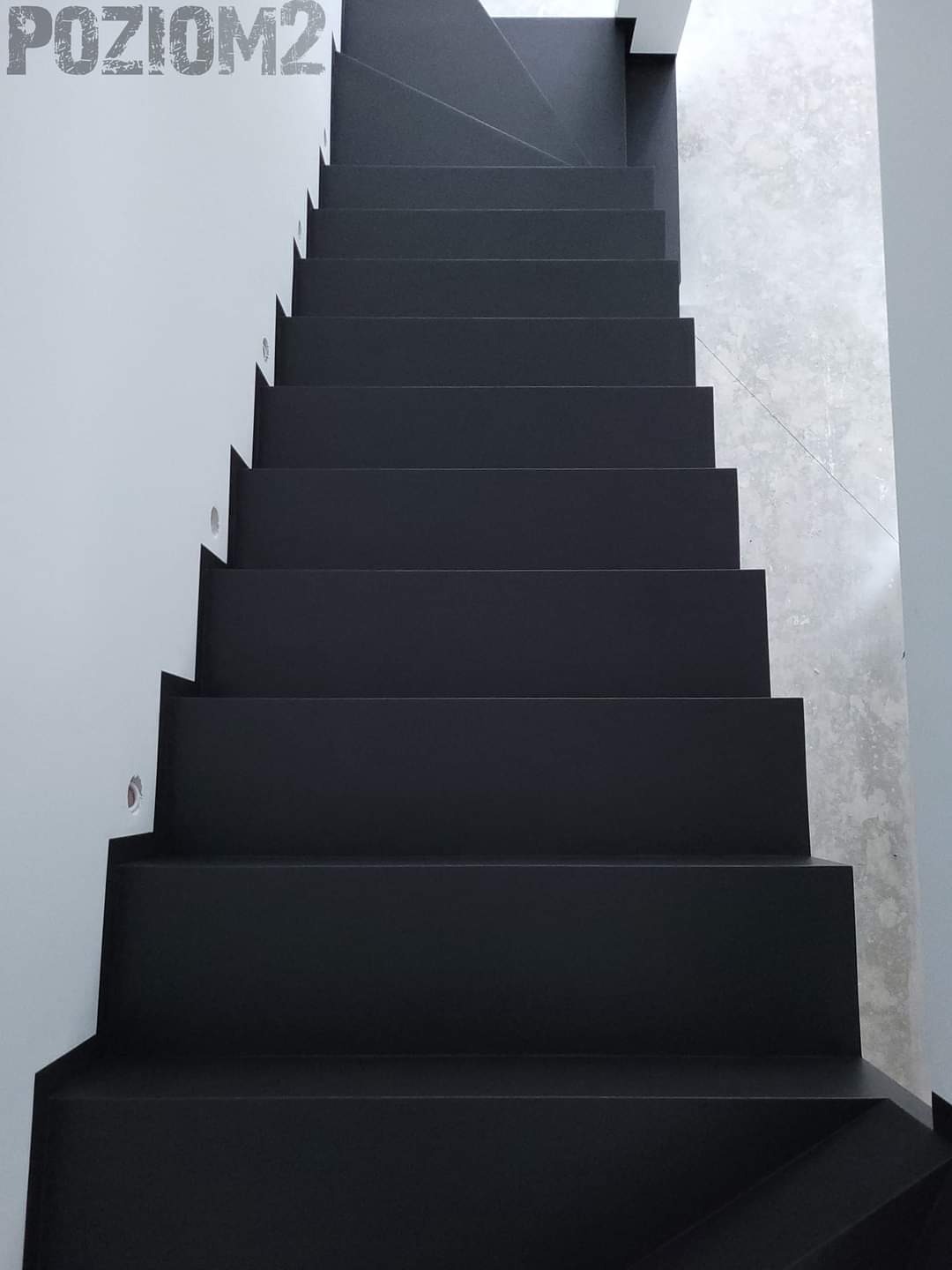Minimalistyczne, czarne schody prowadzące w dół, z subtelnym oświetleniem punktowym w ścianie i surowym betonem na podłodze.