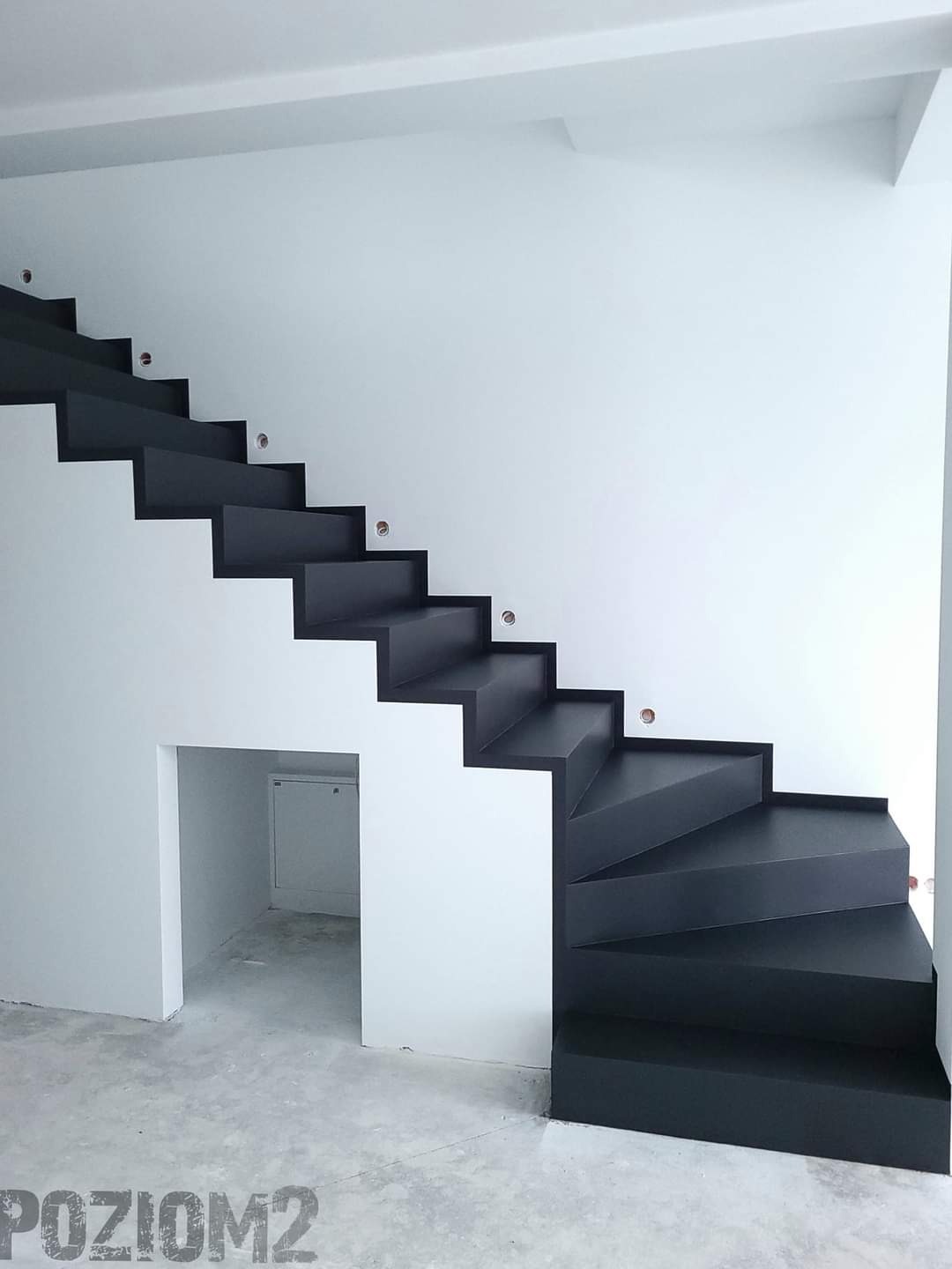 Nowoczesne schody wspornikowe w kolorze antracytowym, z minimalistycznym oświetleniem punktowym w ścianie, prowadzące na wyższą kondygnację, z przestrzenią pod schodami wykorzystaną jako schowek.