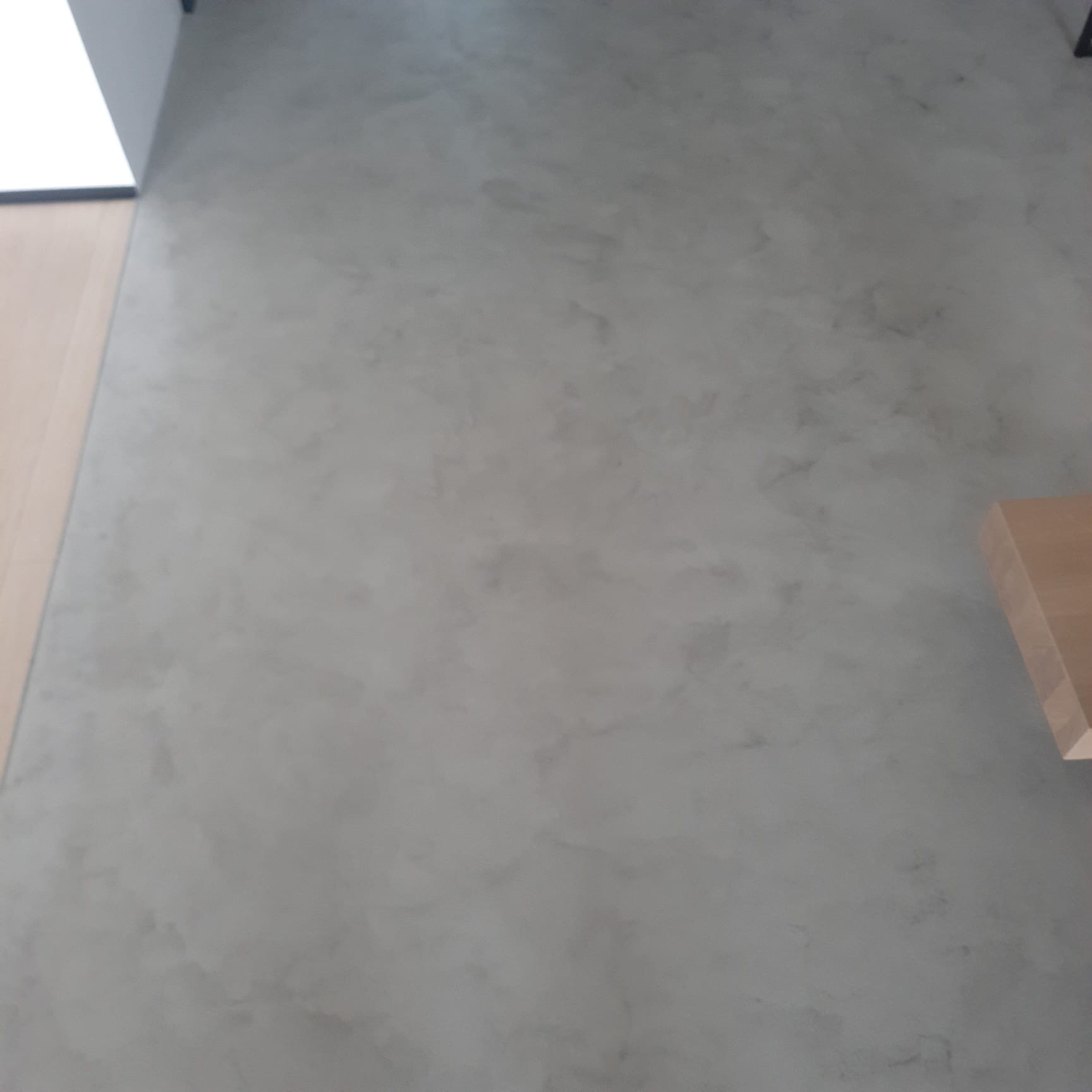 Szara ściana z dekoracyjnym efektem betonu, widoczny fragment drewnianej podłogi i narożnik sufitu.