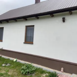 Biała elewacja domu z brązowym dachem i podmurówką, widoczne dwa okna z drewnianymi ramami, numer domu 10, oraz ogródek z roślinami i figurką psa.