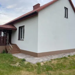 Biały dom jednorodzinny z brązowym dachem i elementami wykończeniowymi, widok z boku na elewację i wejście z werandą, otoczony trawnikiem.