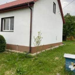 Świeżo otynkowany, biały dom z brązowym dachem i podmur&oacute;wką, widok z boku na trawnik z młodym drzewkiem i pomalowanym ulem w tle.