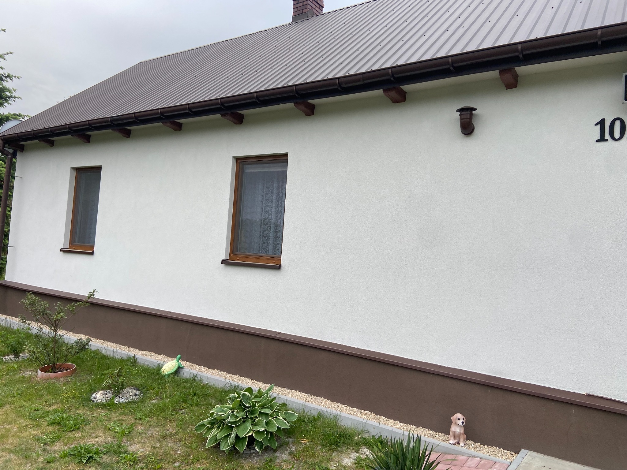Biała elewacja domu z brązowym dachem i podmurówką, widoczne dwa okna z drewnianymi ramami, numer domu 10, oraz ogródek z roślinami i figurką psa.