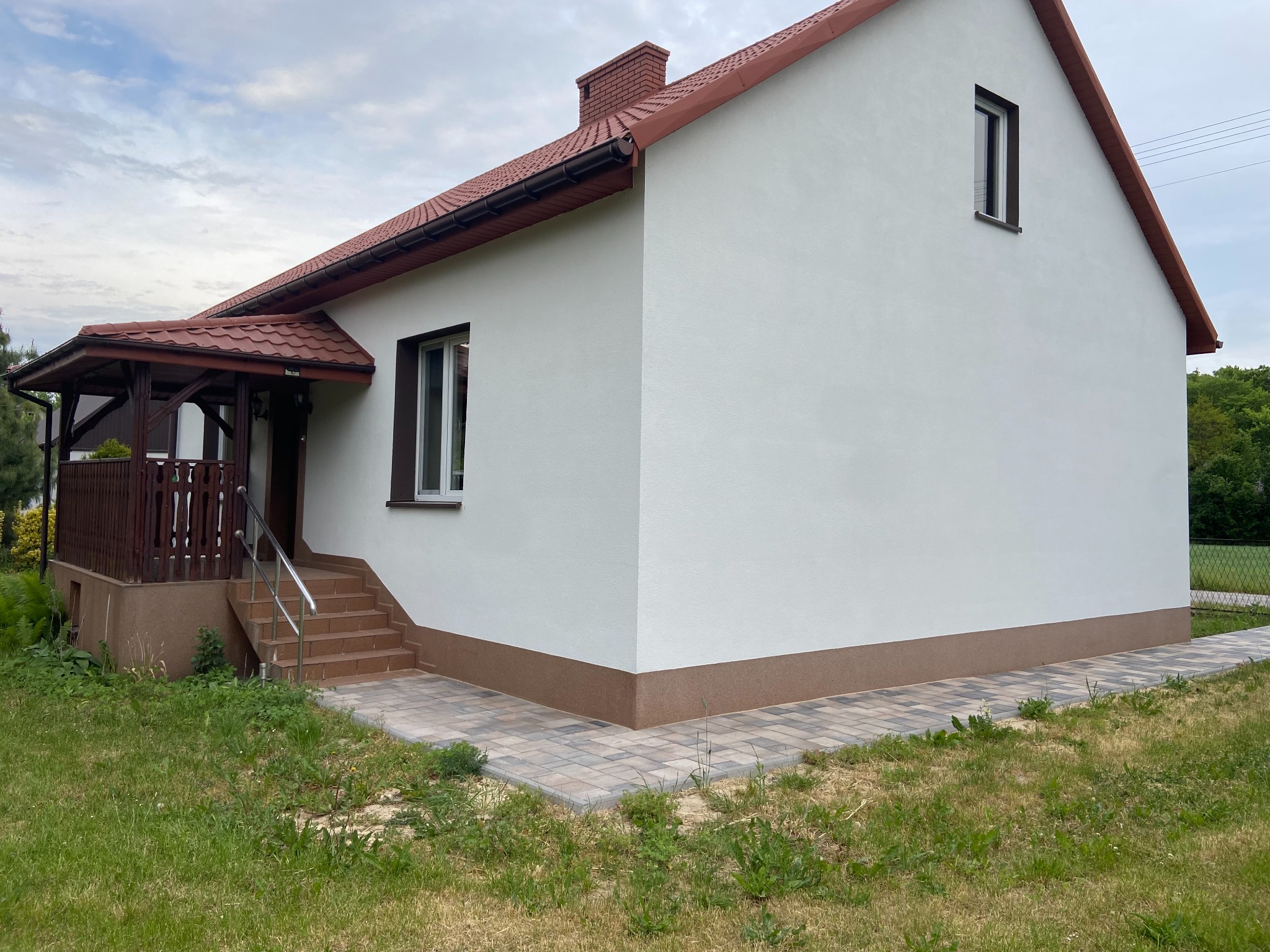 Biały dom jednorodzinny z brązowym dachem i elementami wykończeniowymi, widok z boku na elewację i wejście z werandą, otoczony trawnikiem.