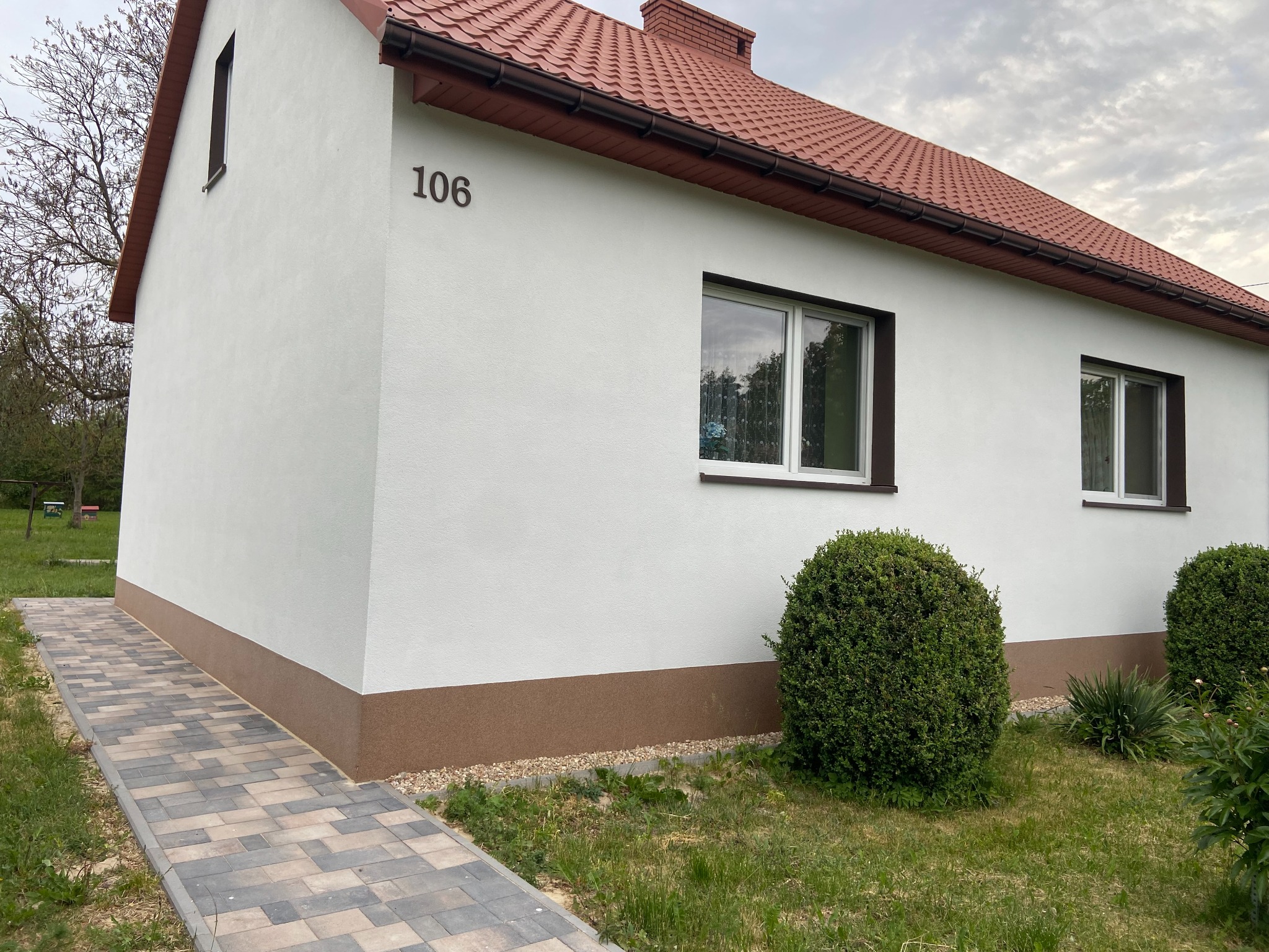 Biały dom jednorodzinny z numerem 106, czerwoną dachówką i brązowym cokołem, otoczony zielenią, z kostką brukową prowadzącą do wejścia.