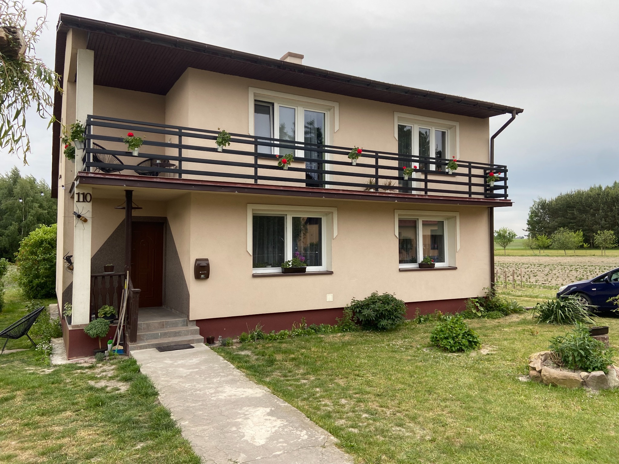 Dwukondygnacyjny dom z beżową elewacją, czarnymi balustradami balkonów z donicami z czerwonymi kwiatami, zadbany trawnik i ścieżka z płyt betonowych prowadząca do wejścia.