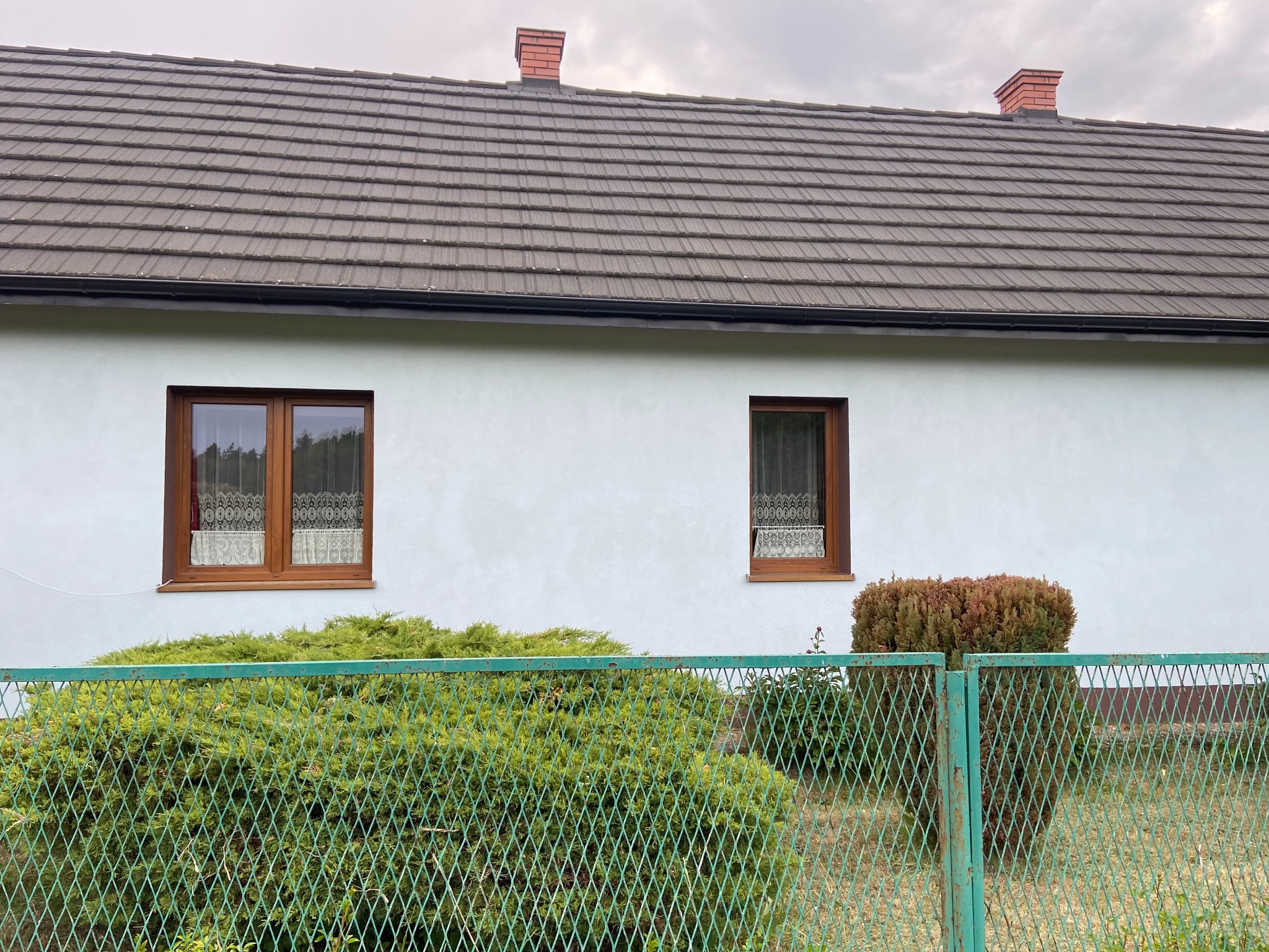 Jasna elewacja domu jednorodzinnego z brązowymi oknami i ciemnym dachem, widoczna zza zielonego metalowego ogrodzenia porośniętego krzewami.