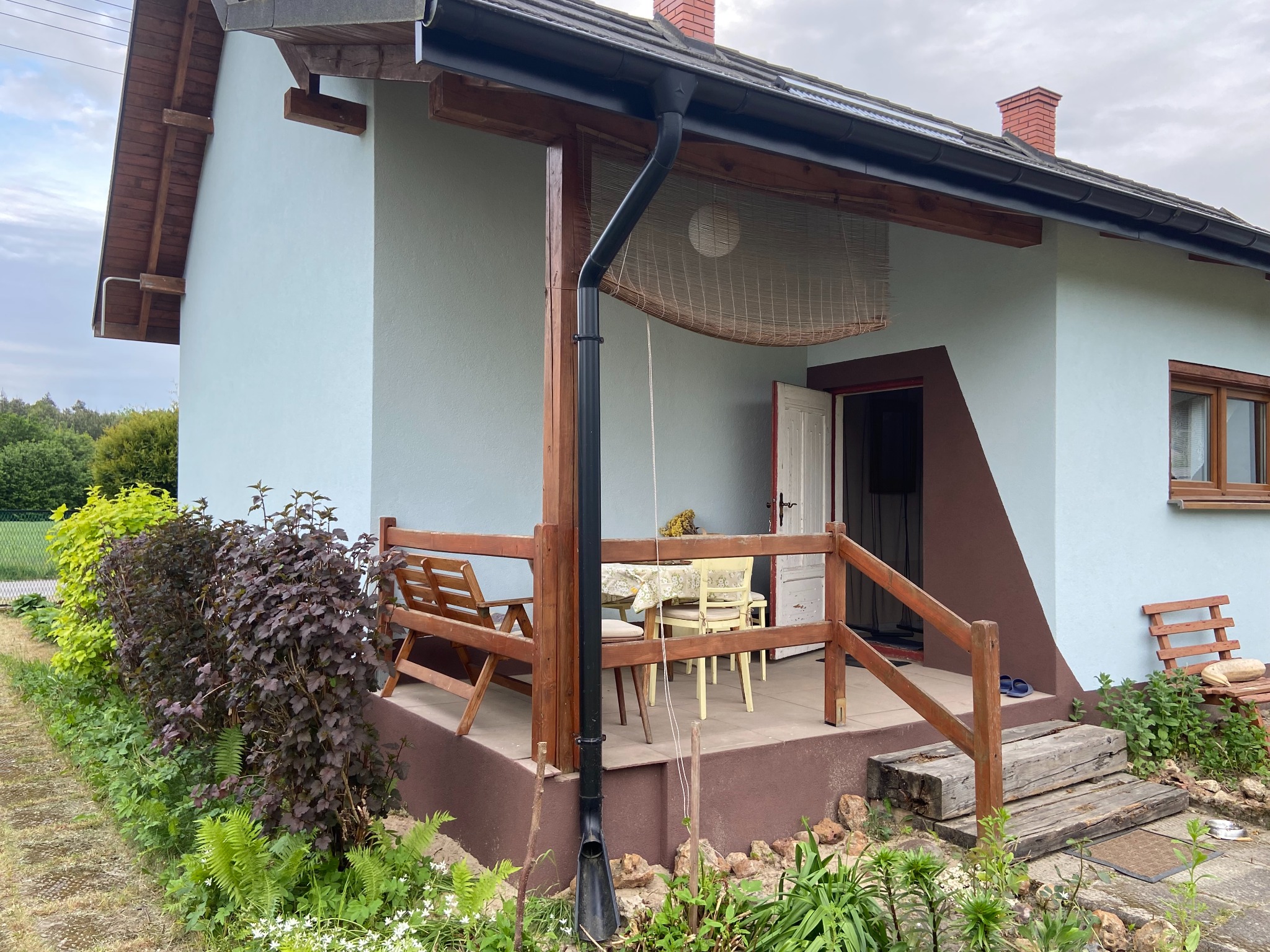 Dom jednorodzinny z jasną elewacją, drewnianym zadaszonym tarasem z meblami ogrodowymi, otoczony zielenią i ozdobnymi krzewami, z widocznym fragmentem rynny.