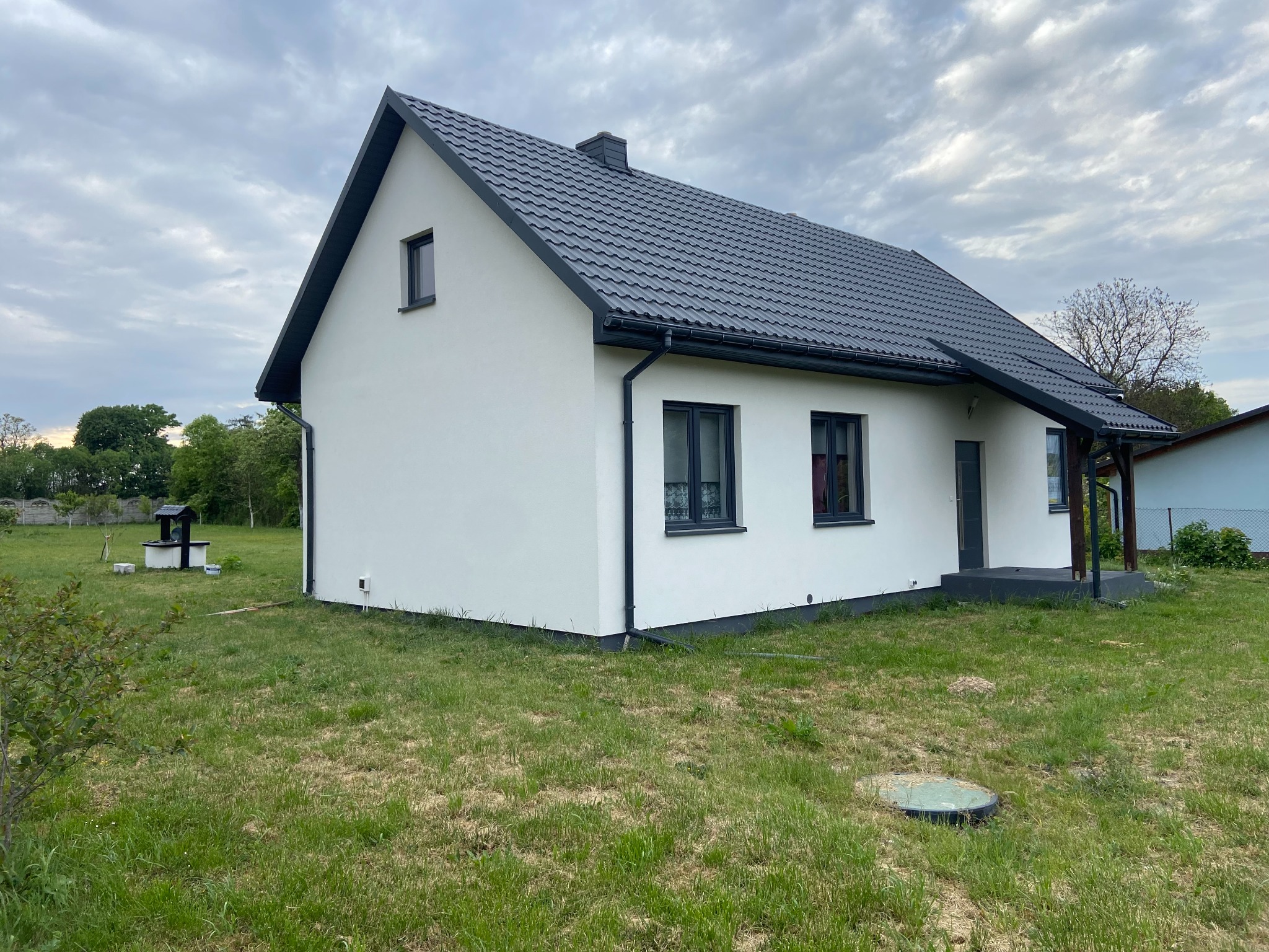 Jednopiętrowy dom z białą elewacją i grafitowym dachem z blachodachówki, widok z boku na trawnik z studnią w tle.