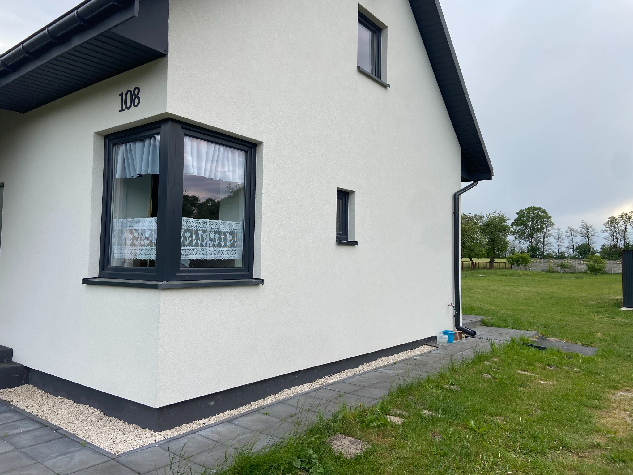 Biała elewacja domu z numerem 108, ciemne obramowania okien i dachu, widok z boku na narożnik budynku, szary chodnik z kostki brukowej i trawnik.