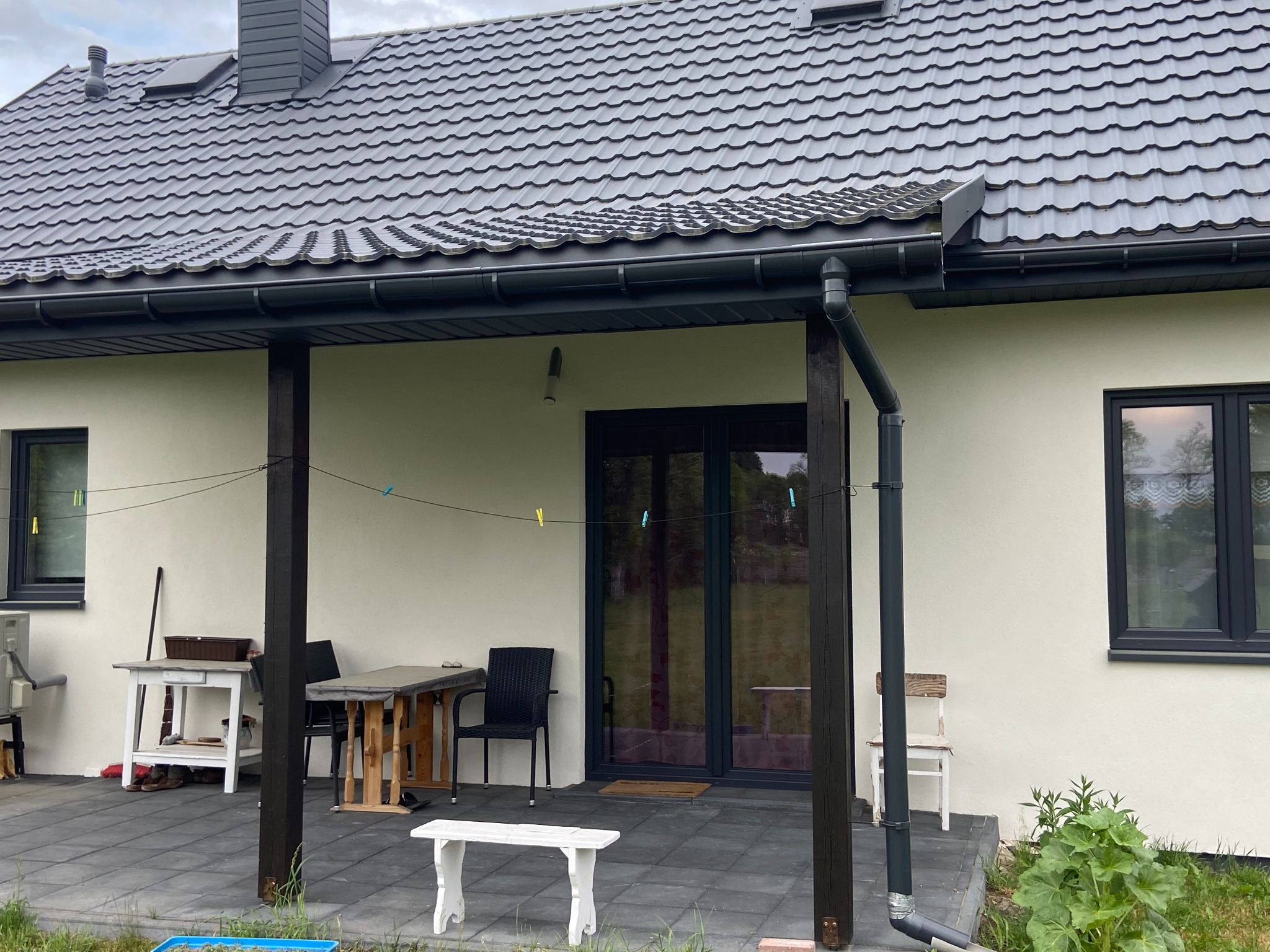 Elewacja domu jednorodzinnego w Nagłowicach po tynkowaniu, z widocznymi czarnymi oknami, ciemną dachówką i elementami drewnianej konstrukcji werandy, na której suszą się ubrania.