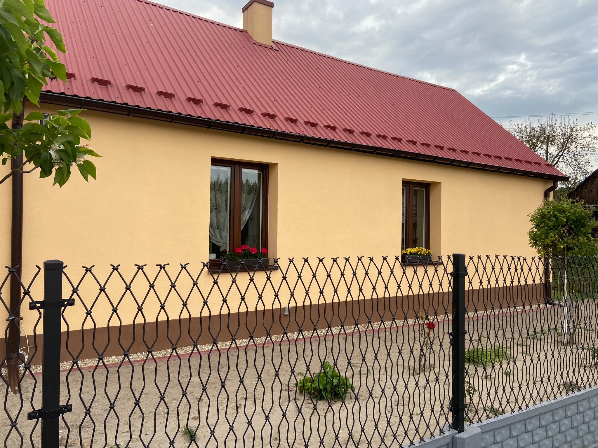 Beżowy dom z czerwonym dachem blachodachówką, ciemnobrązową podmurówką i ramami okiennymi, ozdobiony kwiatami w donicach na parapetach, ogrodzony czarnym, metalowym płotem o falistym wzorze.