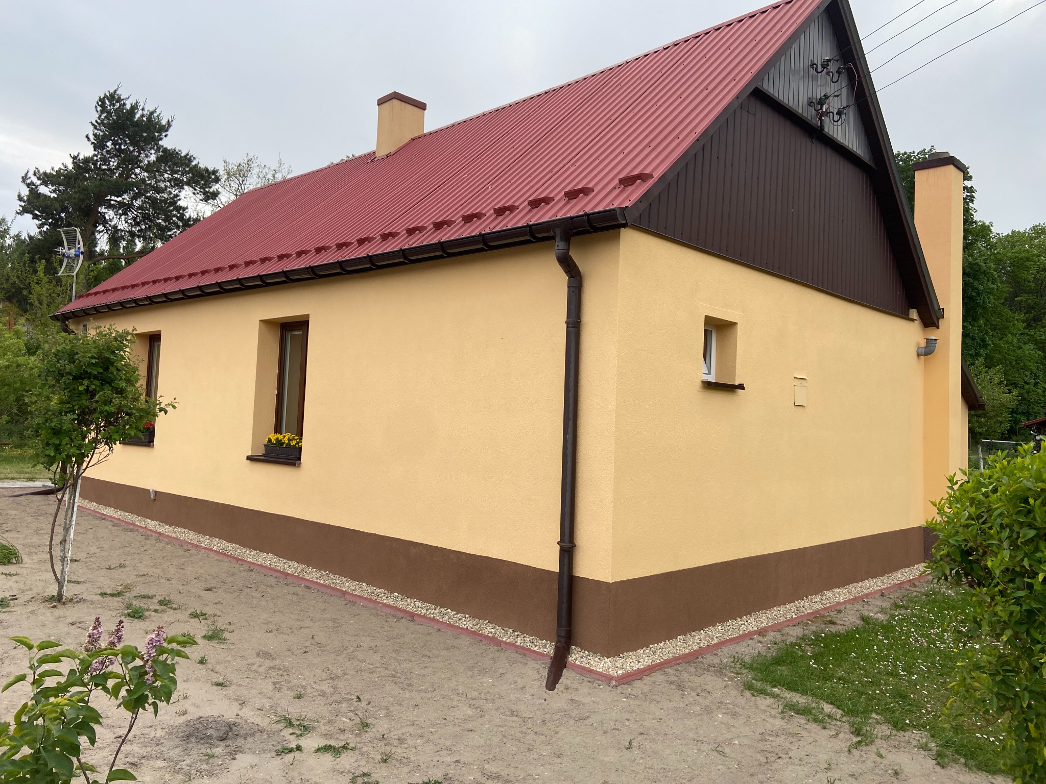 Ukończony dom jednorodzinny z beżową elewacją, brązowym cokołem i czerwoną blachodachówką, widok z boku na niezagospodarowaną działkę.