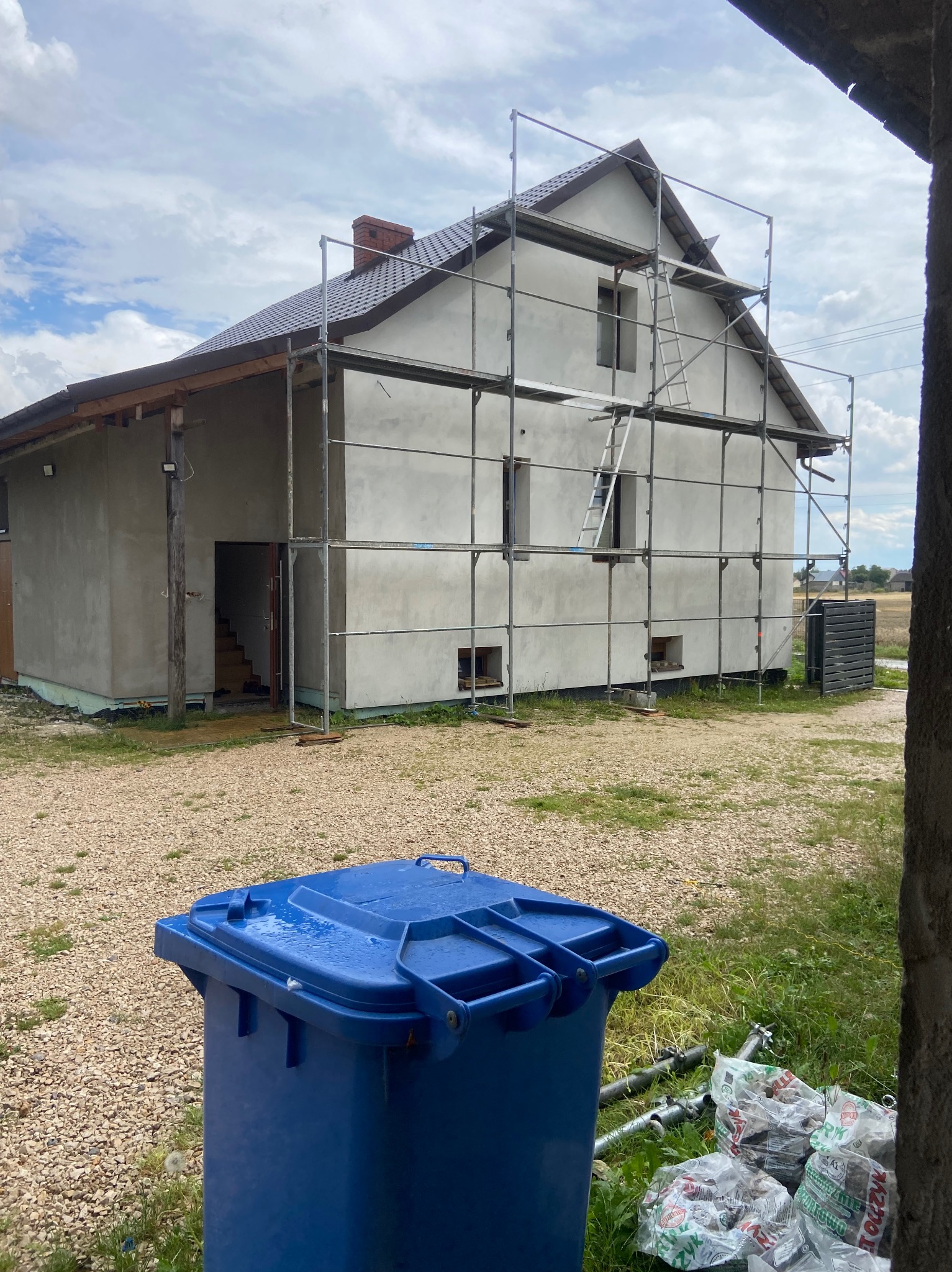 Dom w trakcie tynkowania elewacji, otoczony rusztowaniem. Na pierwszym planie niebieski pojemnik na śmieci i worki z zaprawą. Widoczne elementy krajobrazu.