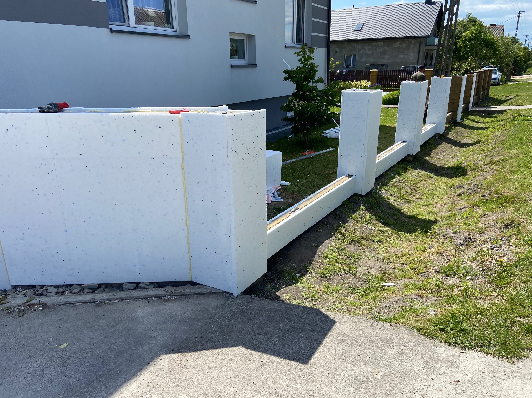 Montaż paneli styropianowych na ogrodzeniu posesji, widoczne elementy mocujące i narzędzia. Prace w toku, jasne oświetlenie dzienne.