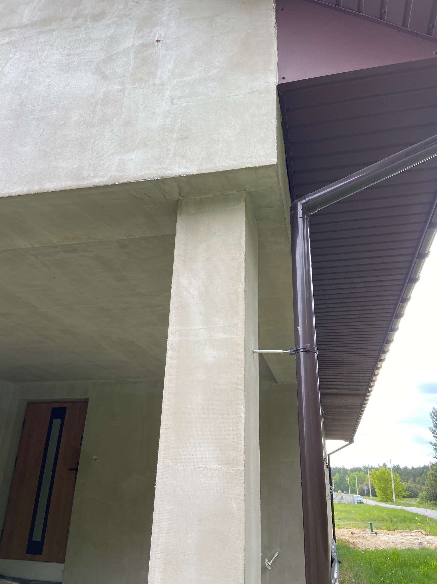 Surowa elewacja domu z widoczną kolumną i orynnowaniem w brązowym kolorze. Świeżo nałożony tynk z widoczną siatką. Widok z dołu na fragment budynku.