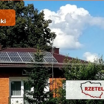 Panele fotowoltaiczne na dachu dwuspadowym z cegły, widoczne z poziomu ogrodu, z fragmentami drzew i błękitnym niebem z chmurami w tle. Dodatkowo widoczny jest znak wodny z napisem 'RZETEL'.