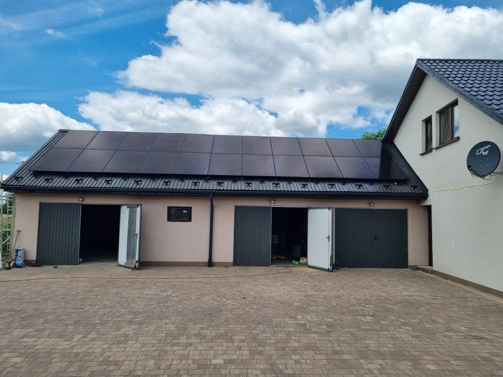 Panele słoneczne zamontowane na dachu budynku gospodarczego z trzema garażami, widoczne błękitne niebo z białymi chmurami.
