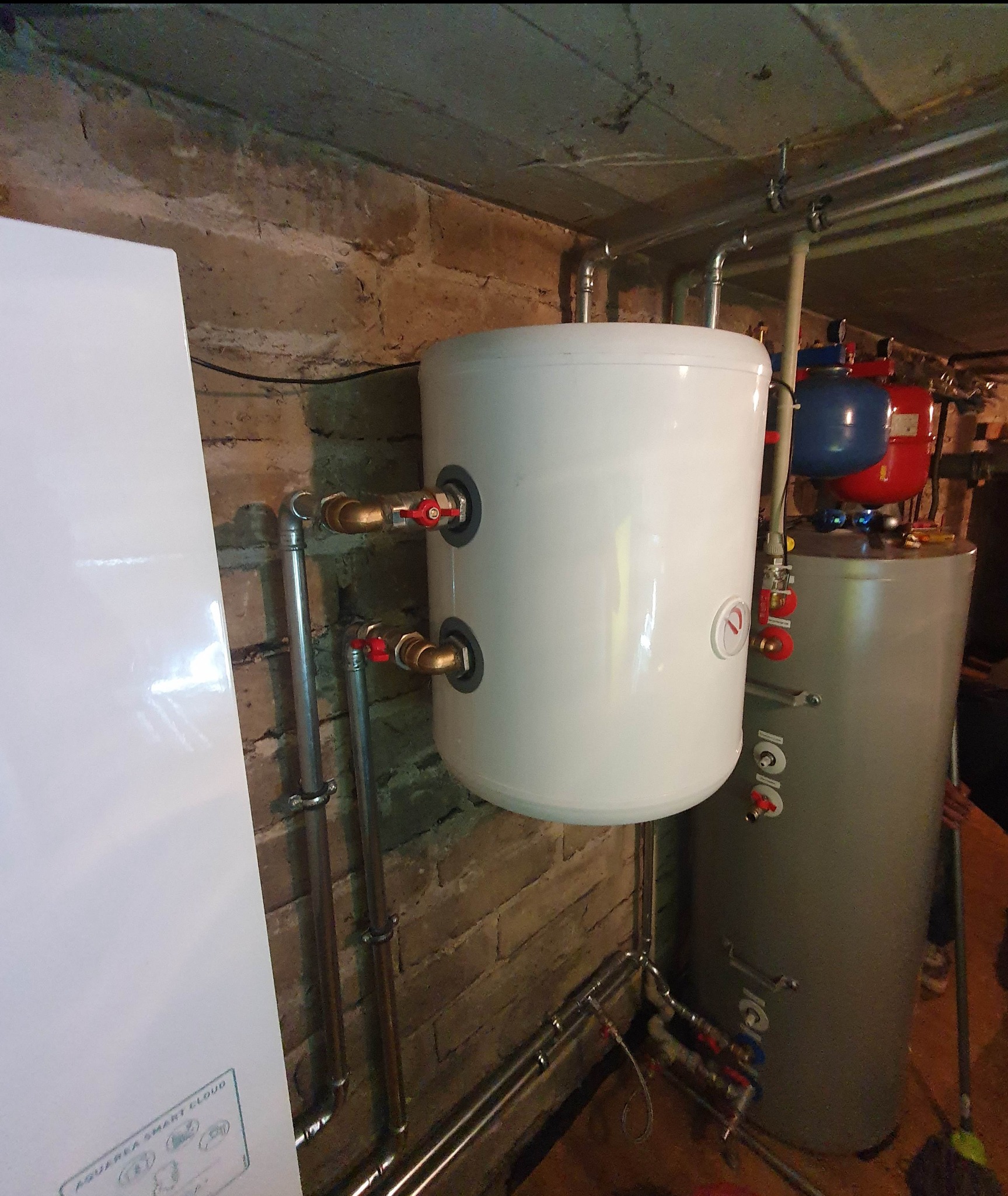 Biały zasobnik ciepłej wody użytkowej zamontowany na ścianie z cegły, widoczne rury miedziane z zaworami, obok szary zbiornik z niebiesko-czerwonym naczyniem wzbiorczym, w tle instalacja rurowa...