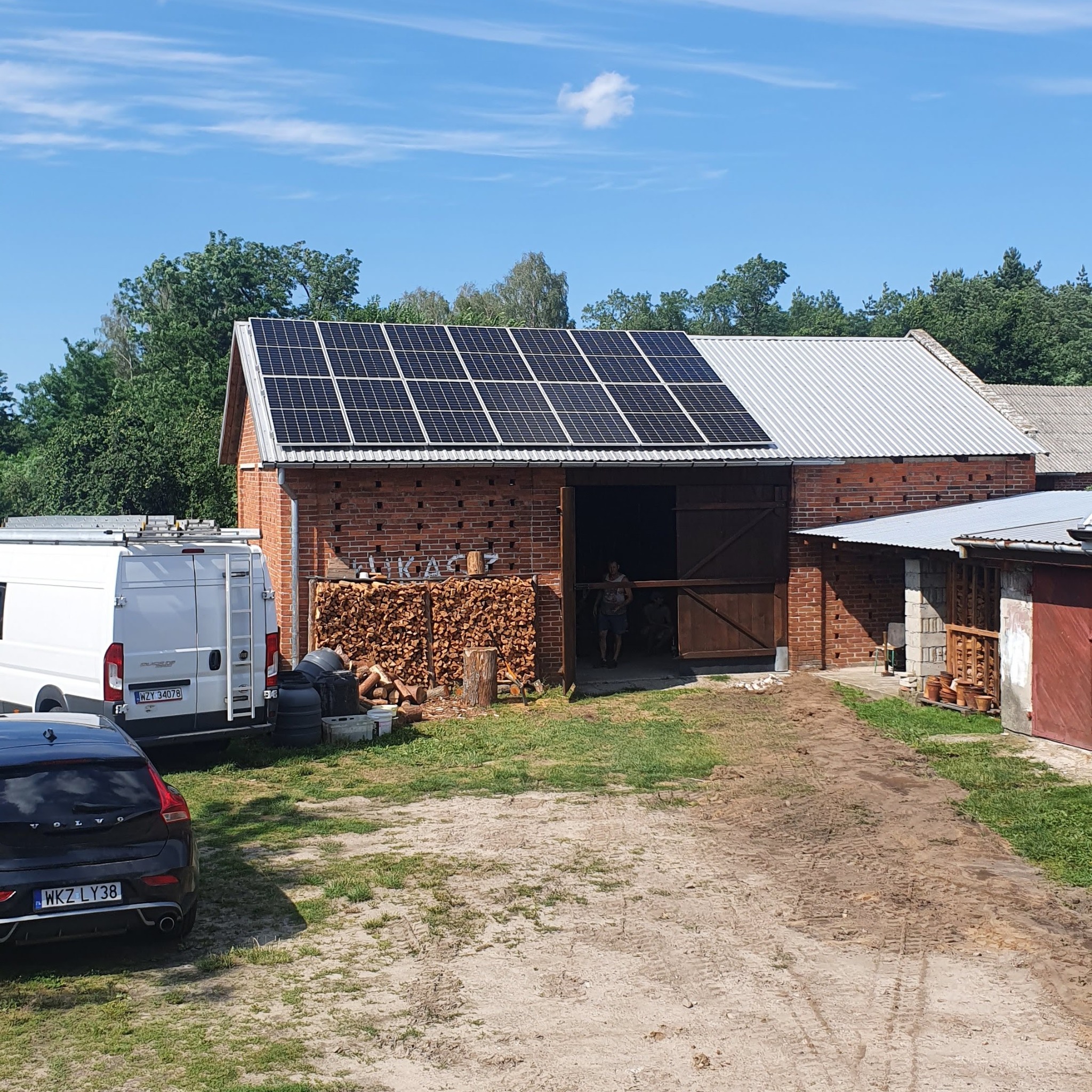 Instalacja paneli fotowoltaicznych na dachu ceglanego budynku gospodarczego w Kozienicach. Na pierwszym planie zaparkowane samochody i sterta drewna.