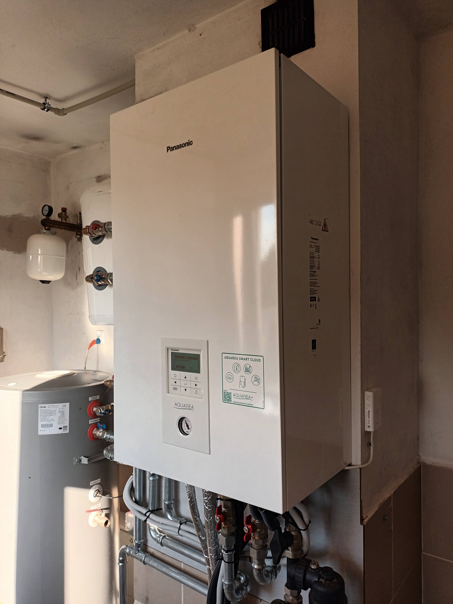 Biała pompa ciepła Panasonic Aquarea zamontowana w kotłowni, widoczne rury i zbiornik buforowy Ferroli, instalacja połączona z systemem Aquarea Smart Cloud.