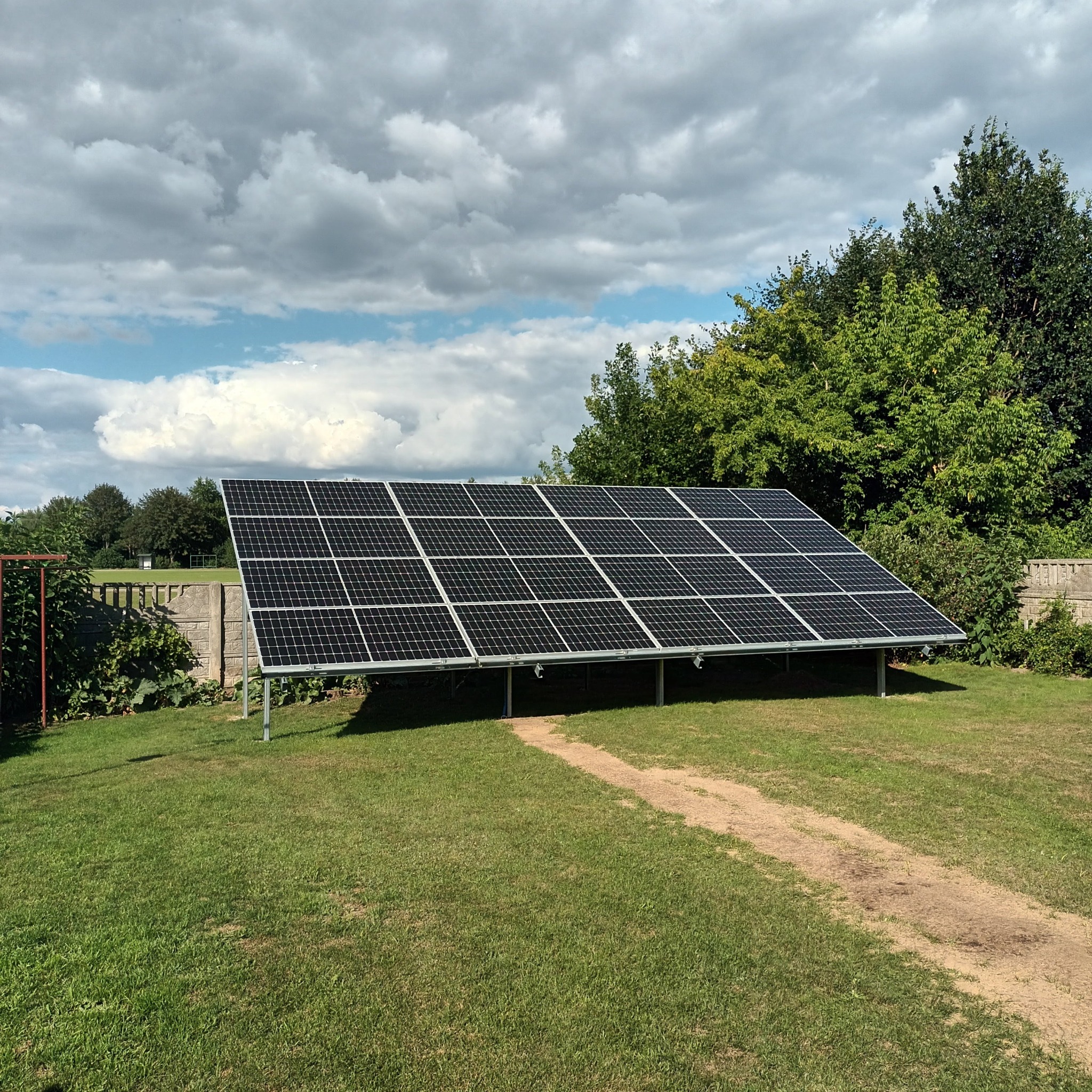 Panele fotowoltaiczne ustawione na metalowej konstrukcji na trawniku przed domem jednorodzinnym, widoczne drzewa i pochmurne niebo.