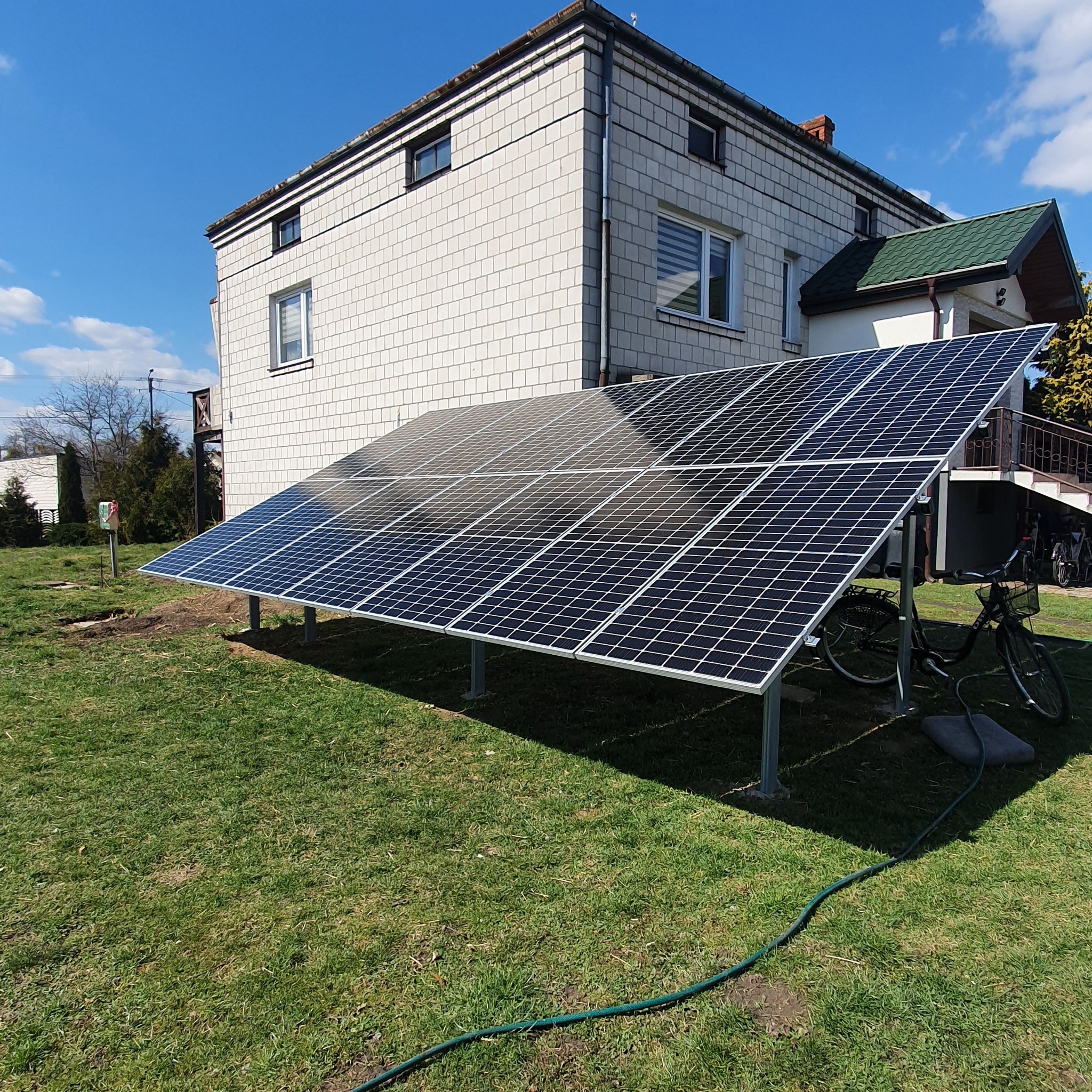 Panel fotowoltaiczny na trawniku przed domem z jasnej cegły, rower oparty o konstrukcję paneli, widoczny wąż ogrodowy na trawie.