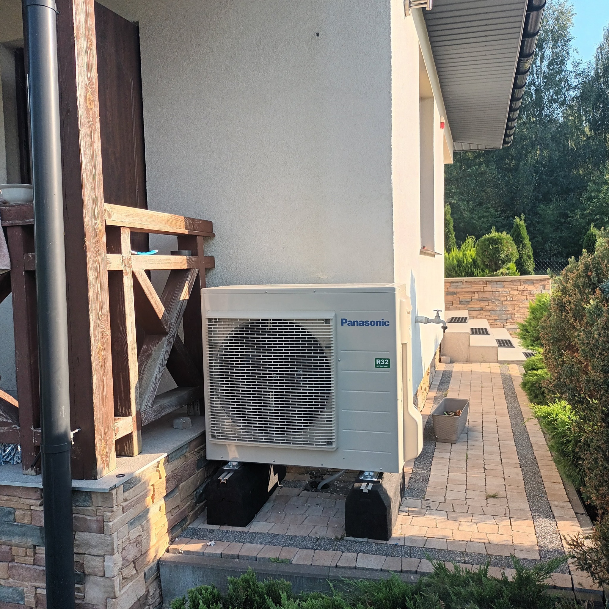 Zewnętrzna jednostka klimatyzacji Panasonic R32 zamontowana na czarnych, antywibracyjnych podstawach na kostce brukowej, obok drewnianej konstrukcji werandy i ścieżki w ogrodzie.