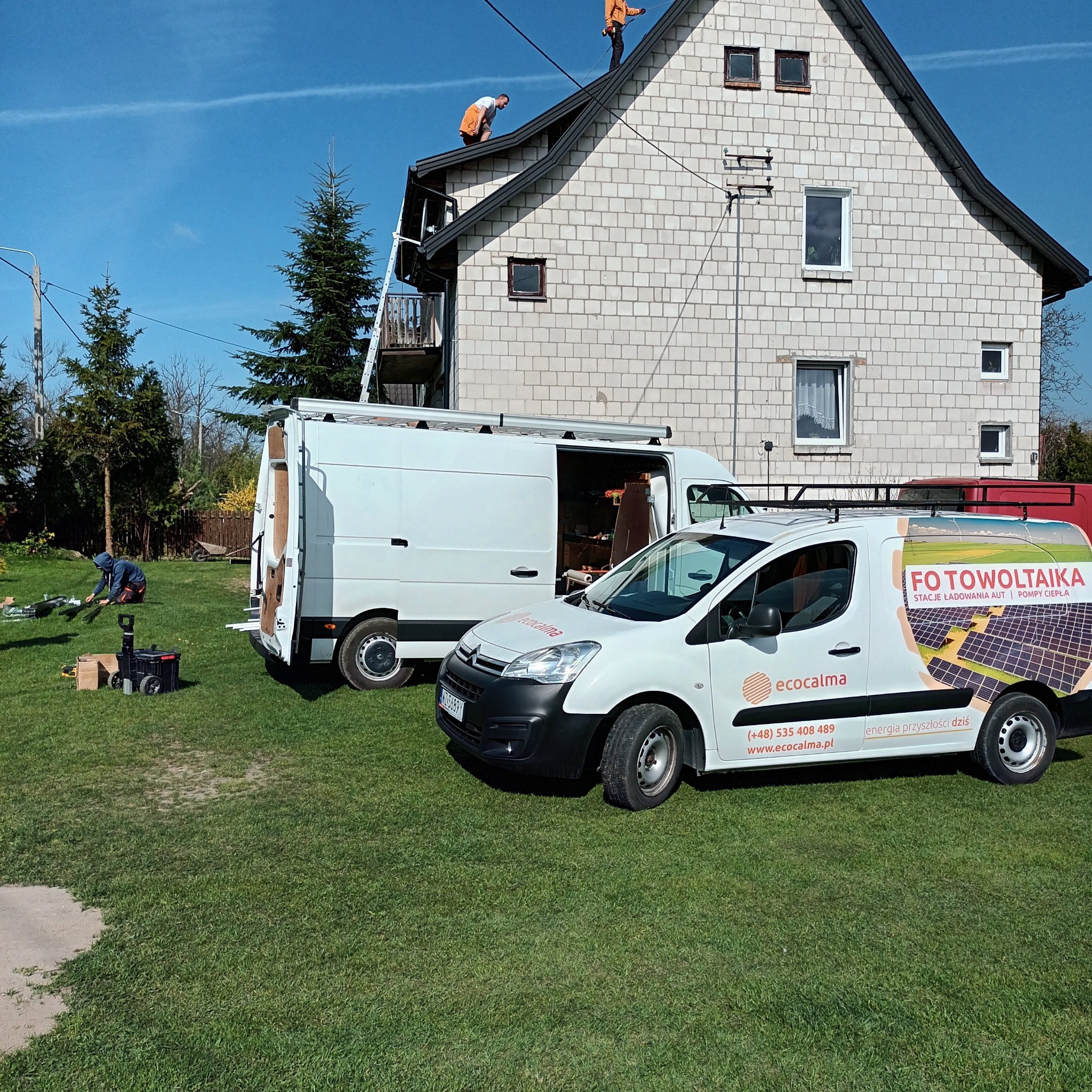 Montaż paneli na dachu domu jednorodzinnego z dwoma samochodami dostawczymi firmy instalacyjnej na trawniku. Na samochodzie widoczne logo firmy i napis 'Fotowoltaika, stacje ładowania aut, pompy...
