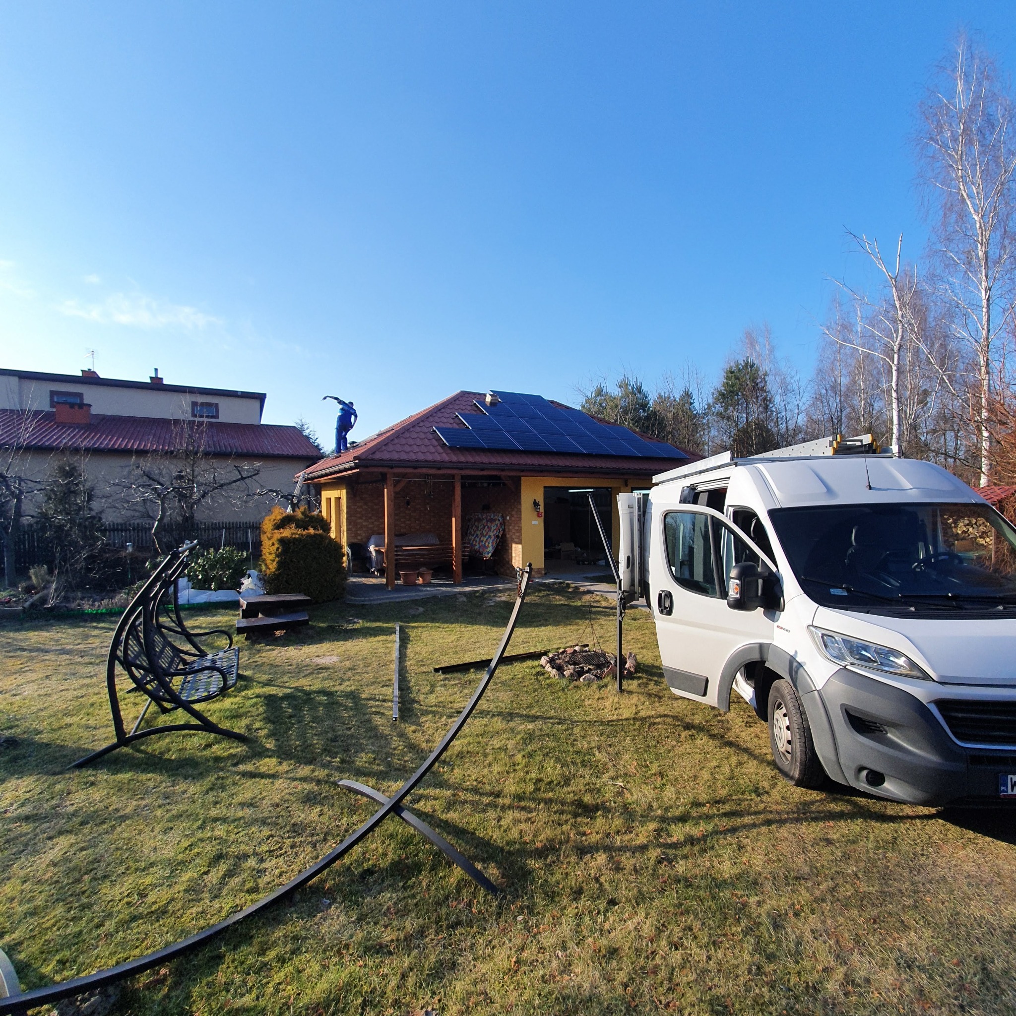 Instalacja paneli fotowoltaicznych na dachu domu jednorodzinnego, widoczny monter w niebieskim kombinezonie, biały samochód dostawczy z otwartymi drzwiami i rozłożona drabina, słoneczny dzień.
