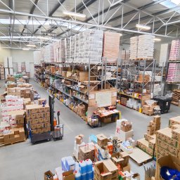 PRIMA - Chemia gospodarcza - Magazyn z wysokimi regałami wypełnionymi kartonami i opakowaniami różnych produktów, w tym płynów w niebieskich butelkach, widoczne wózki transportowe i stanowisko pracy z mapą na ścianie.