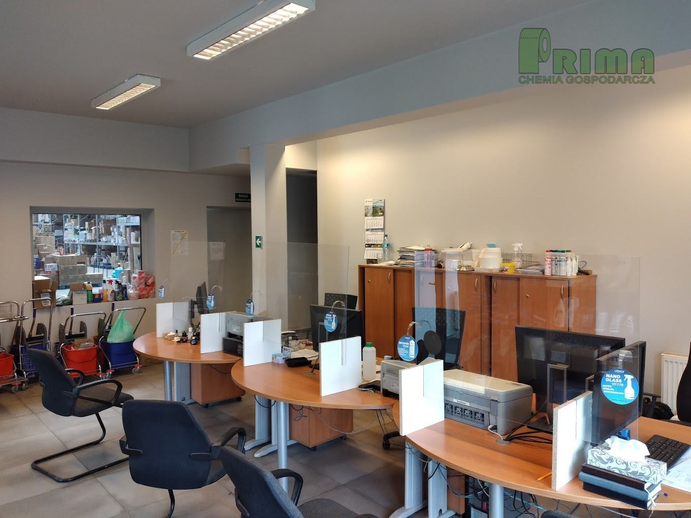 Biuro z zabezpieczeniami z pleksi na biurkach, widoczne butelki z płynami dezynfekcyjnymi i w tle magazyn z artykułami chemii gospodarczej.