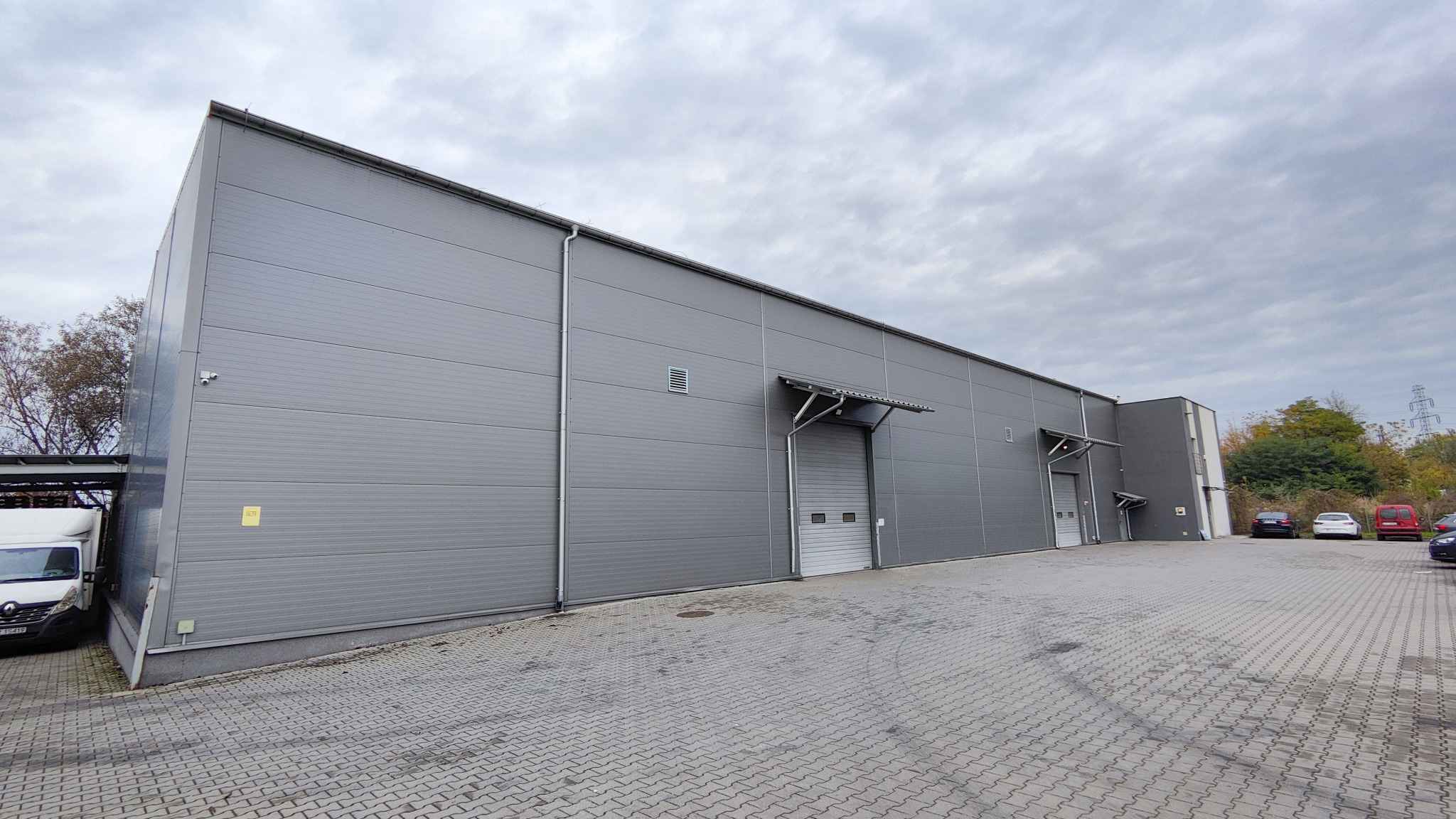 Szary budynek magazynowy z blachy trapezowej, widok z zewnątrz na plac manewrowy z kostki brukowej, pochmurne niebo.