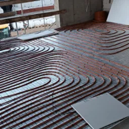 Montaż ogrzewania podłogowego: pomarańczowe rury rozłożone na metalowej siatce w surowym wnętrzu budynku w trakcie budowy, widoczne płyty izolacyjne i zwój rur.